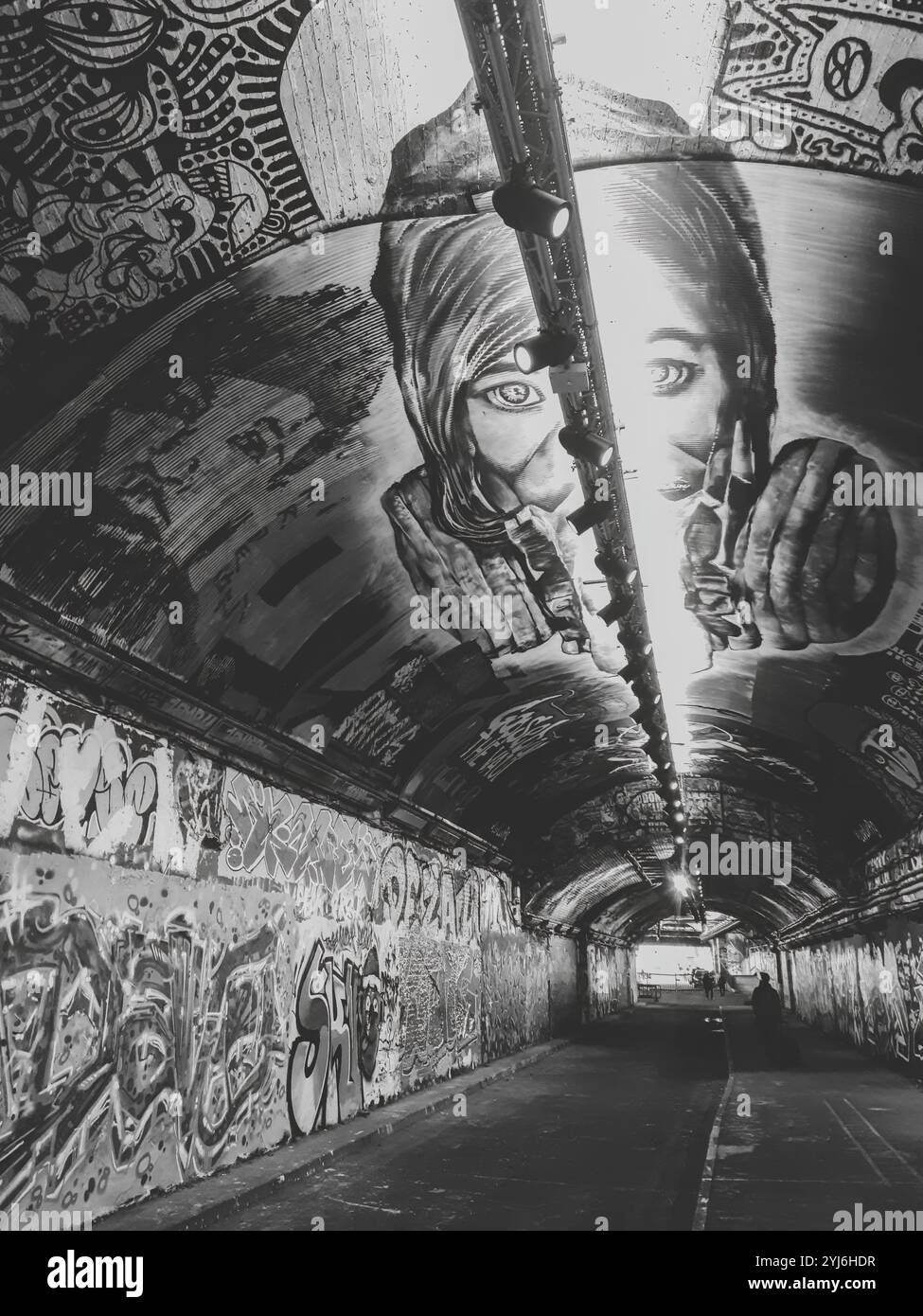 Schwarzweiß-Foto eines mit Graffiti bedeckten Tunnels mit dramatischen Kunstwerken an der Decke, die eine Person mit einem Schal und einem intensiven Blick darstellen. Stockfoto