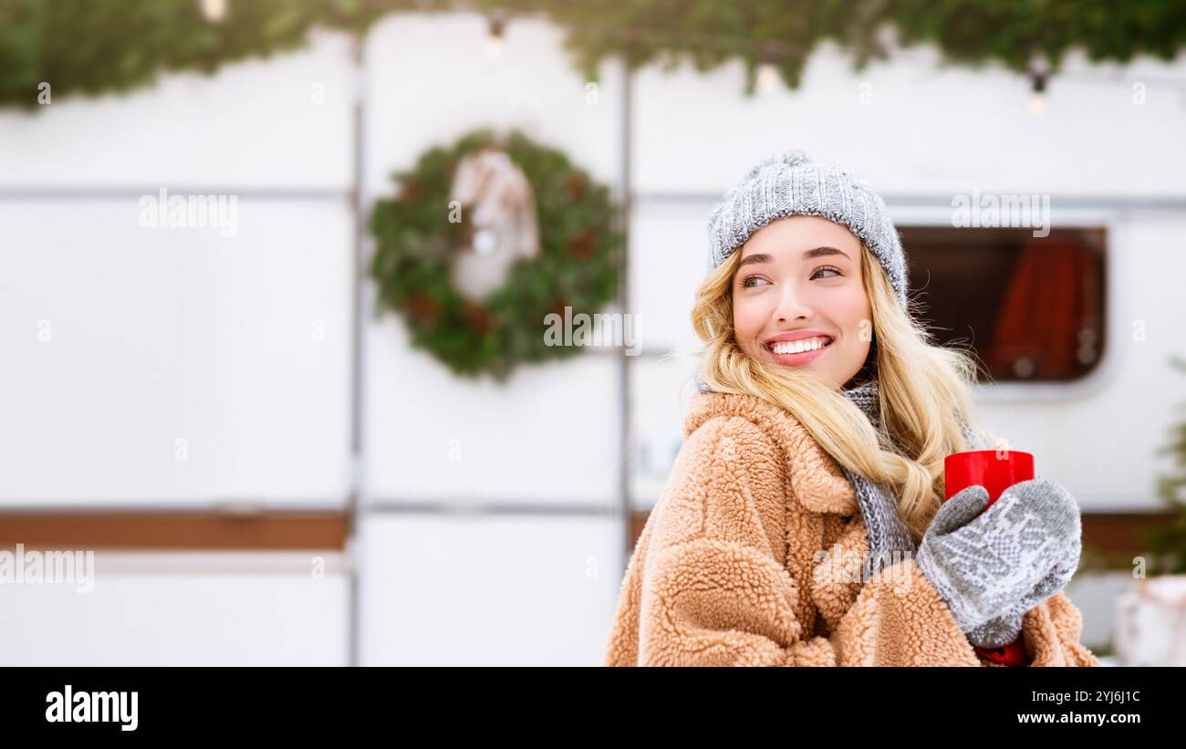 Eine junge Frau, die strahlend lächelt, hält ein warmes Getränk in gemütlicher Winterkleidung. Sie steht in der Nähe der Weihnachtsdekoration und fängt das Wesen ein Stockfoto