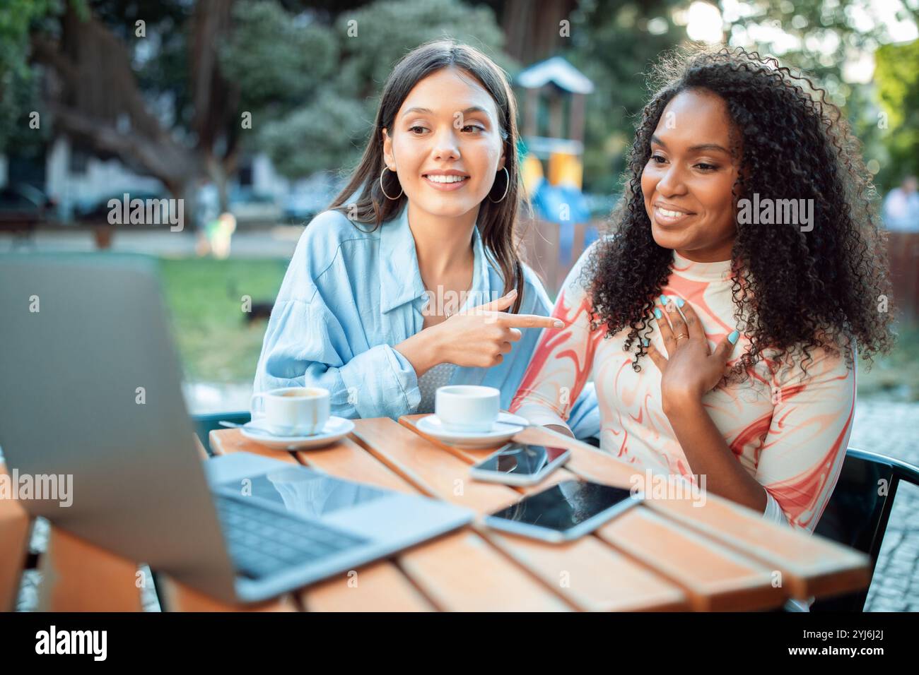 Zwei Freunde sitzen an einem Holztisch in einem lebhaften Café, lächeln und unterhalten sich, während sie einen Kaffee genießen. Laptops und Telefone sind um sie herum und schaffen eine entspannte Atmosphäre Stockfoto