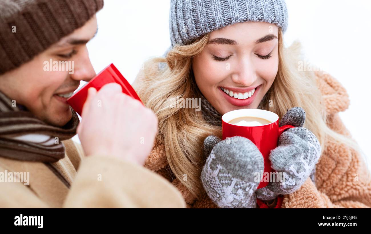 Ein junges Paar isst warme Getränke, während es in einer verschneiten Landschaft lächelt. Gebündelt in gemütliche Winterkleidung teilen sie einen freudigen Moment inmitten der Kälte des Th Stockfoto