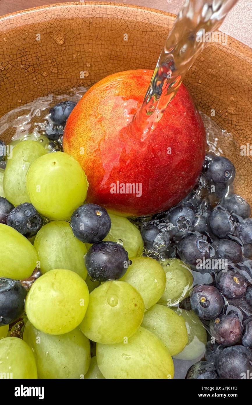 Nahaufnahme von frischem Obst, Heidelbeeren, Pfirsich und Trauben in einer Schüssel unter fließendem Wasser, während die Früchte gewaschen werden. Stockfoto