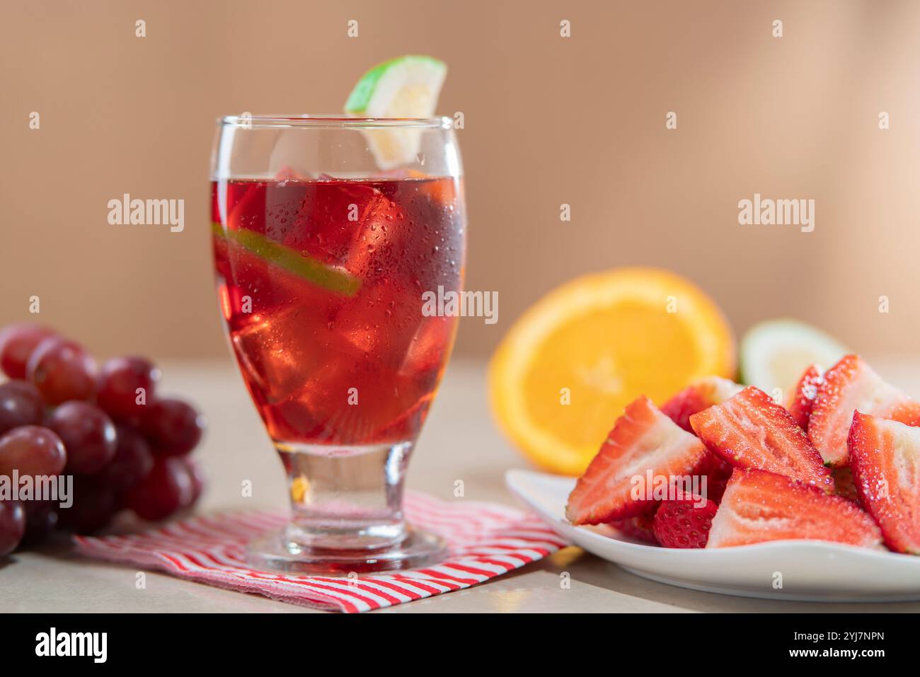 Nahaufnahme eines Glasbechers mit hausgemachtem Sommerwein im Vordergrund neben einem Teller mit gehackten Erdbeeren vor hellbraunem Hintergrund Stockfoto