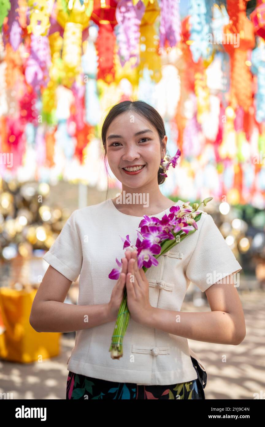 Eine charmante asiatische Frau in traditionellem nordischem Kleid steht in einem Tempel, lächelt in die Kamera und hält einen Blumenstrauß aus Orchideen mit farbenfroher Lanze Stockfoto
