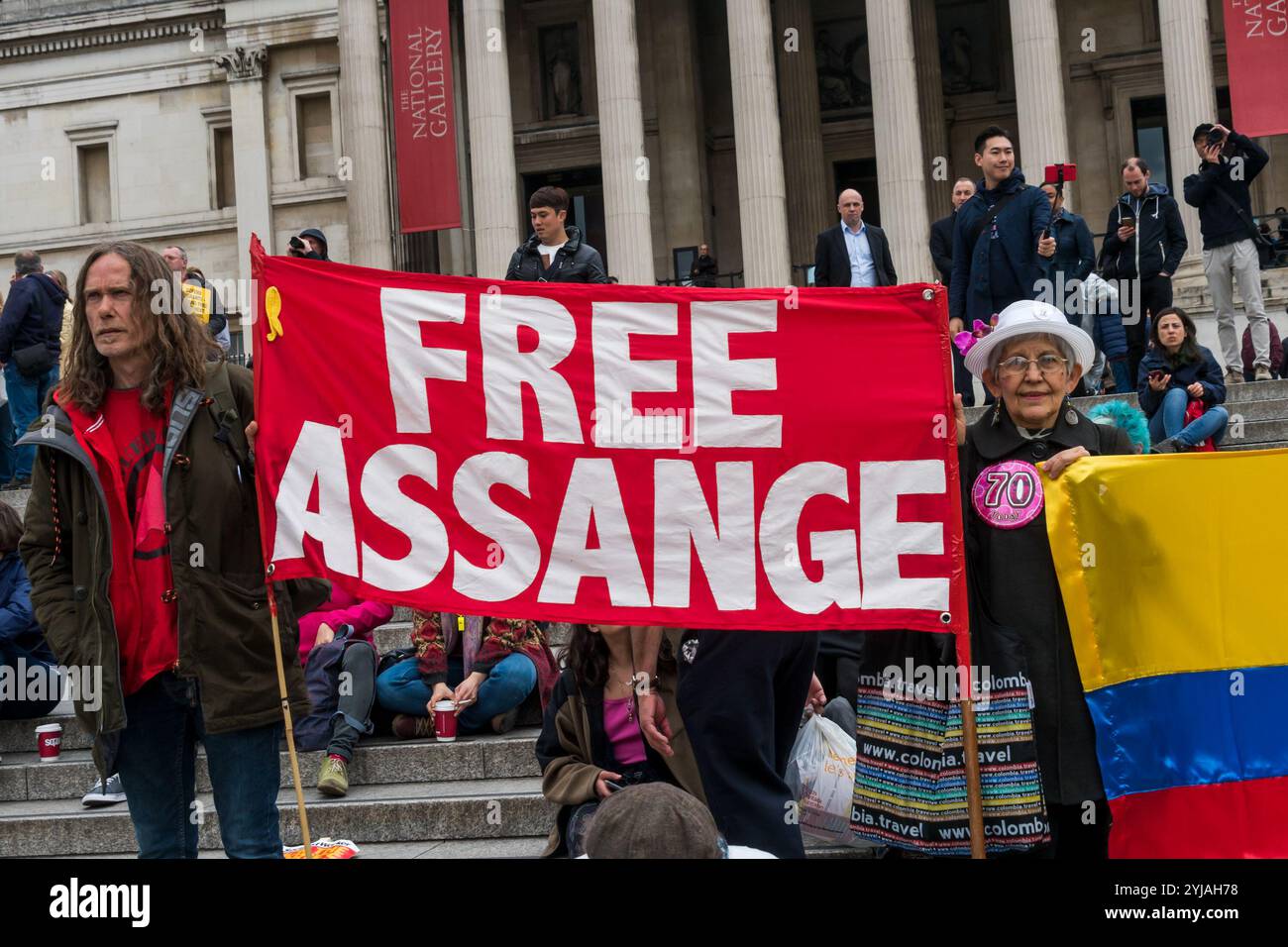 London, Großbritannien. Mai 2018. Menschen, darunter eine Frau, die ein Abzeichen zu ihrem 70. Geburtstag trägt, halten kolumbianische Fahnen und ein Free Assange Banner auf den Stufen des Trafalgar Square bei der Kundgebung, darunter viele aus Londons internationalen und Migrantengemeinschaften, die den Internationalen Arbeitstag feiern. Einige Gewerkschafter und Aktivisten hielten Reden und schlossen ein kurzes Schweigen zum Gedenken an Mehmet Aksoy ein, der in Syrien während der Dreharbeiten mit kurdischen Kämpfern getötet wurde und bei früheren Ereignissen für die Kurden gesprochen hatte. Am Ende der Kundgebung gab es eine Rede von Brixton ritzy Gewerkschafter Kelly Stockfoto