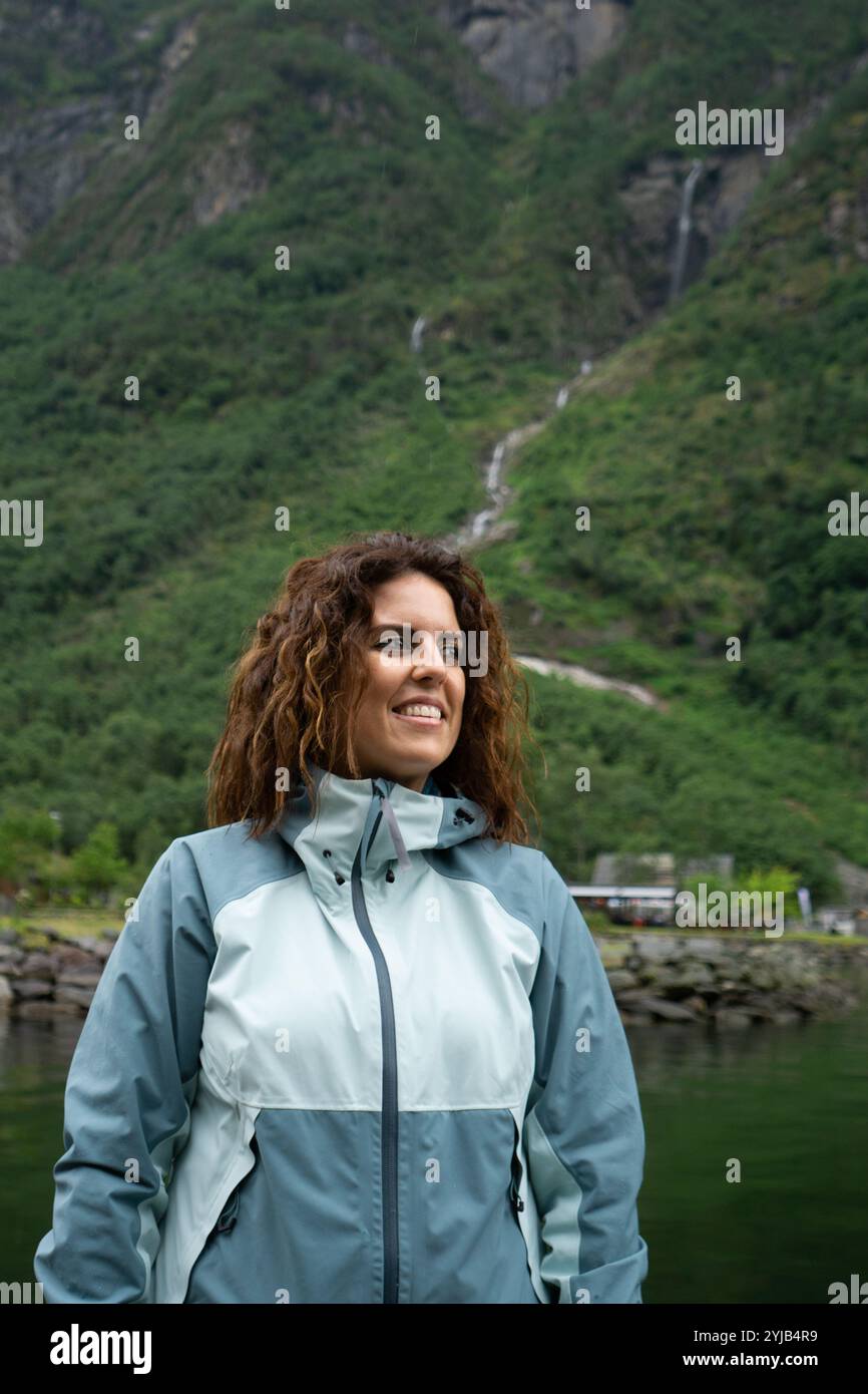 Eine junge Brünette im Alter von 30 Jahren mit Regenjacke blickt zur Seite, mit einer üppig grünen Berglandschaft und einem kaskadierenden Wasserfall im Hintergrund Stockfoto