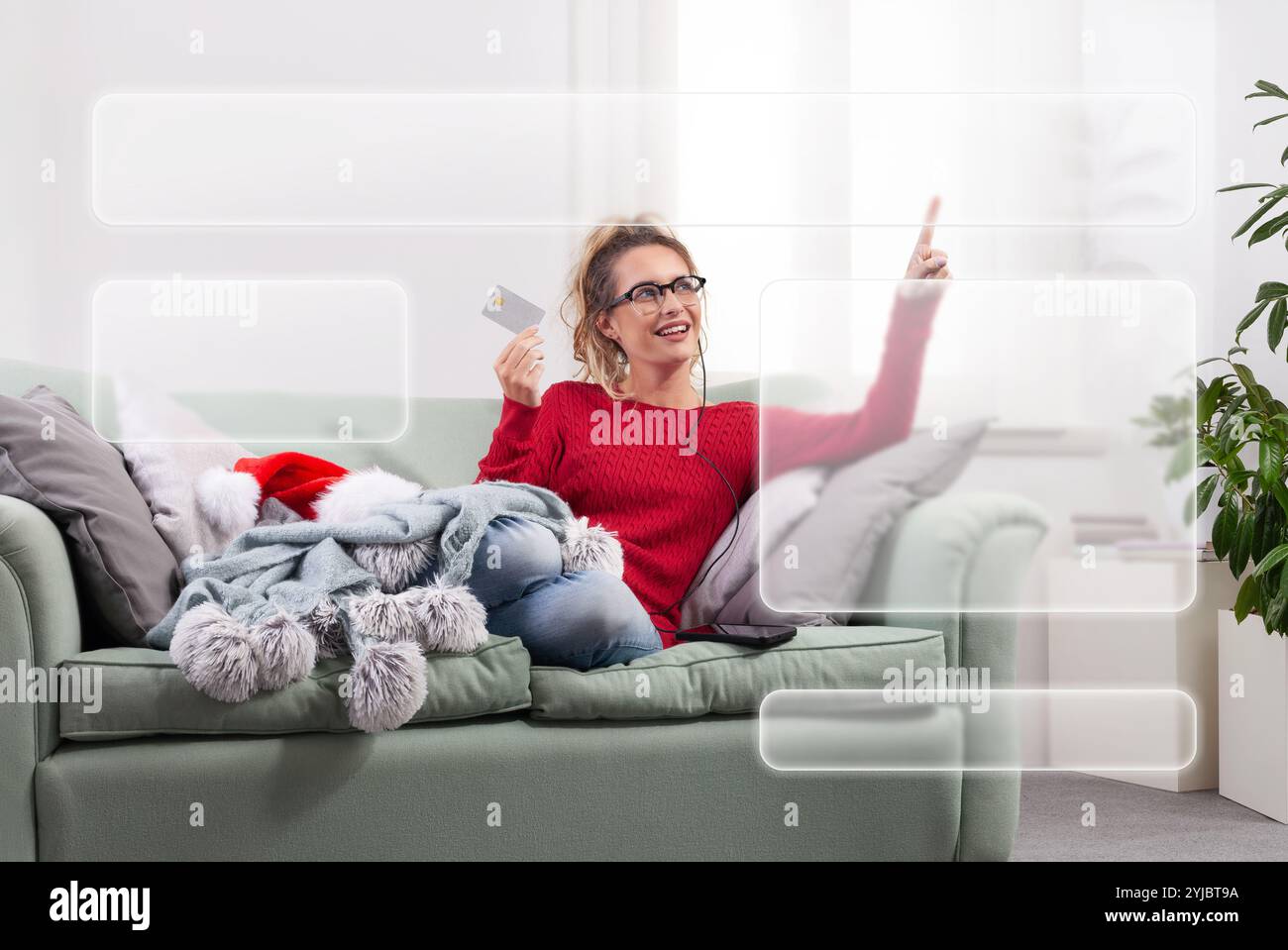 Eine Frau sitzt zu Hause bequem auf ihrer Couch, nutzt ihre intelligente Brille, um im Internet nach Weihnachtseinkäufen zu stöbern, während sie ihre Kreditkarte verwendet Stockfoto