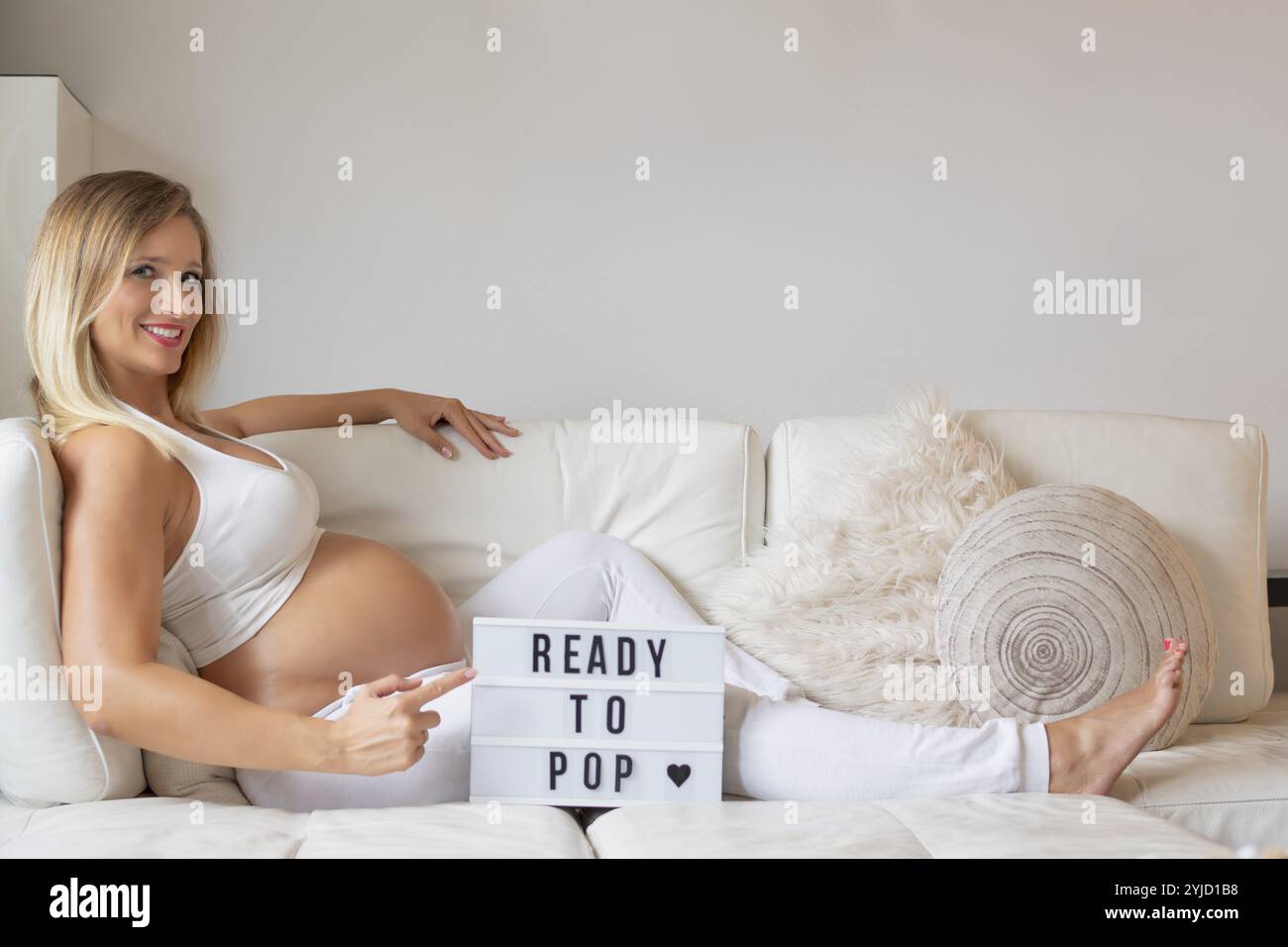 Junge Frau, blond, schwanger, 9 Monate, auf Sofa liegend, er zeigt auf das Schild READY TO POP, Smiling, Stuttgart, Baden-Württemberg, Deutschland, Europa Stockfoto