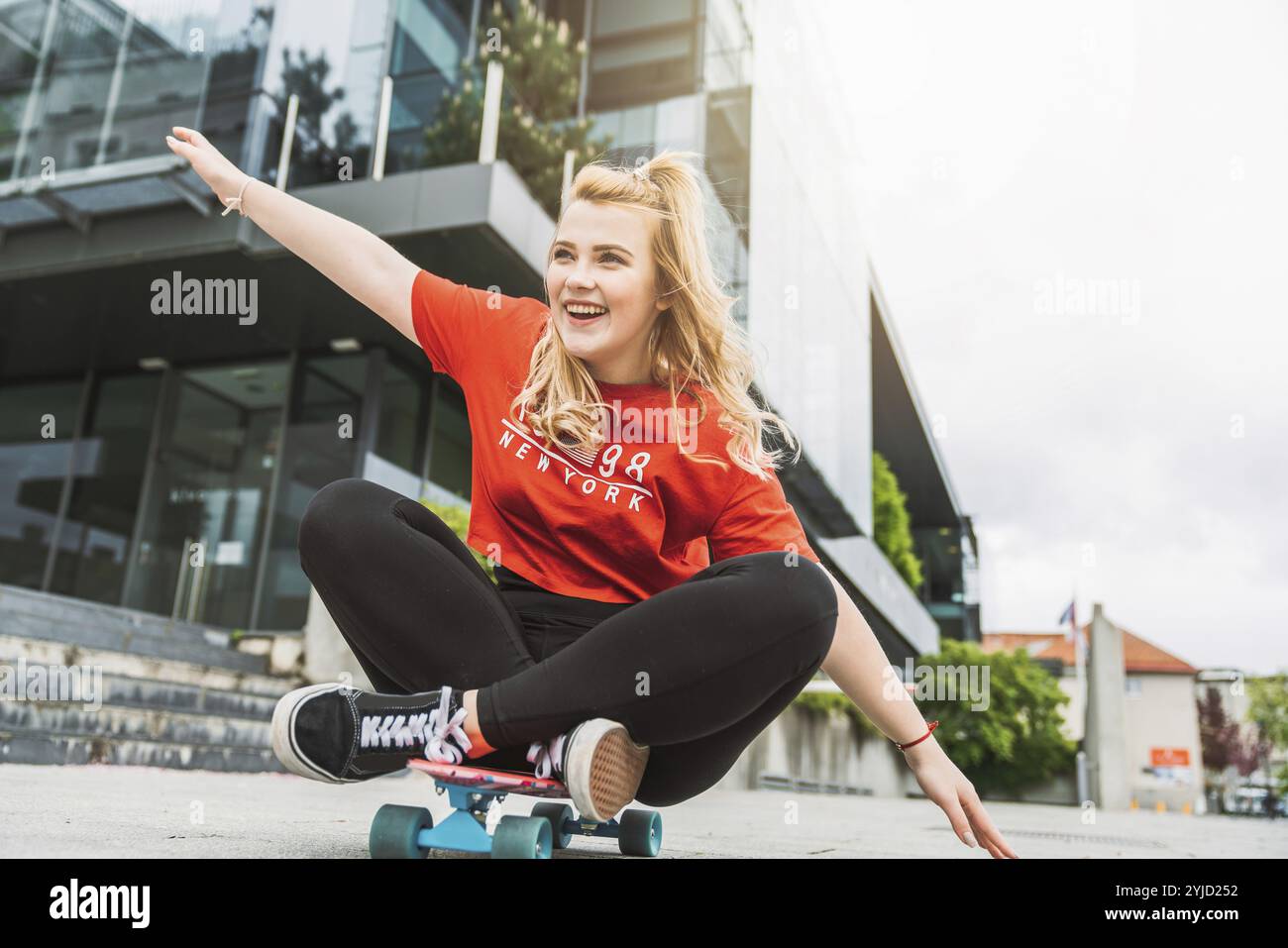 Junge blonde weiße Mädchen, die draußen Spaß auf einem Skateboard hat, schwarze Leggings und rotes Hemd, Sonnenlicht. Teenager lacht, hat draußen Spaß Stockfoto