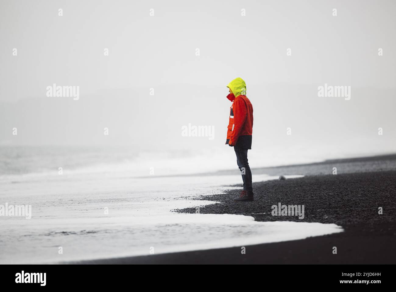 Der Mann in wasserdichter Jacke und Hose, der am Strand mit schwarzem vulkanischem Sand steht, Reynisfjara, südlich von Island. Isländische Natur, Atlantik Stockfoto