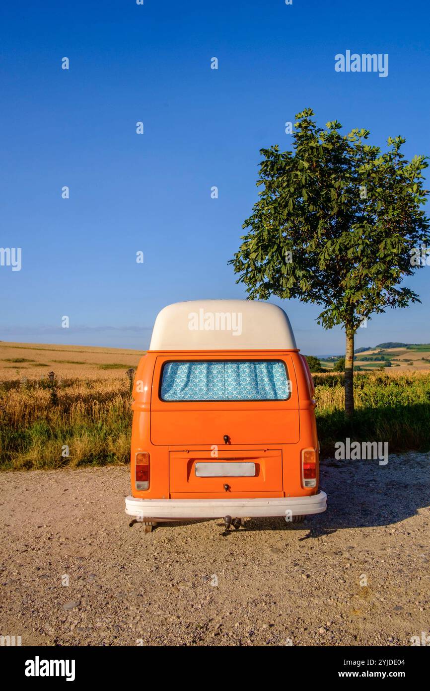 Ein Orange-weisser VW Bus Bulli steht auf einem Schotterparkplatz an einem Sommermorgen, Baden Württemberg, Deutschland. Ein Bulli steht in der Landschaft *** ein orange-weißer VW-Bus steht an einem Sommermorgen auf einem Schotterparkplatz, Baden-Württemberg, Deutschland Ein Van steht in der Landschaft Stockfoto