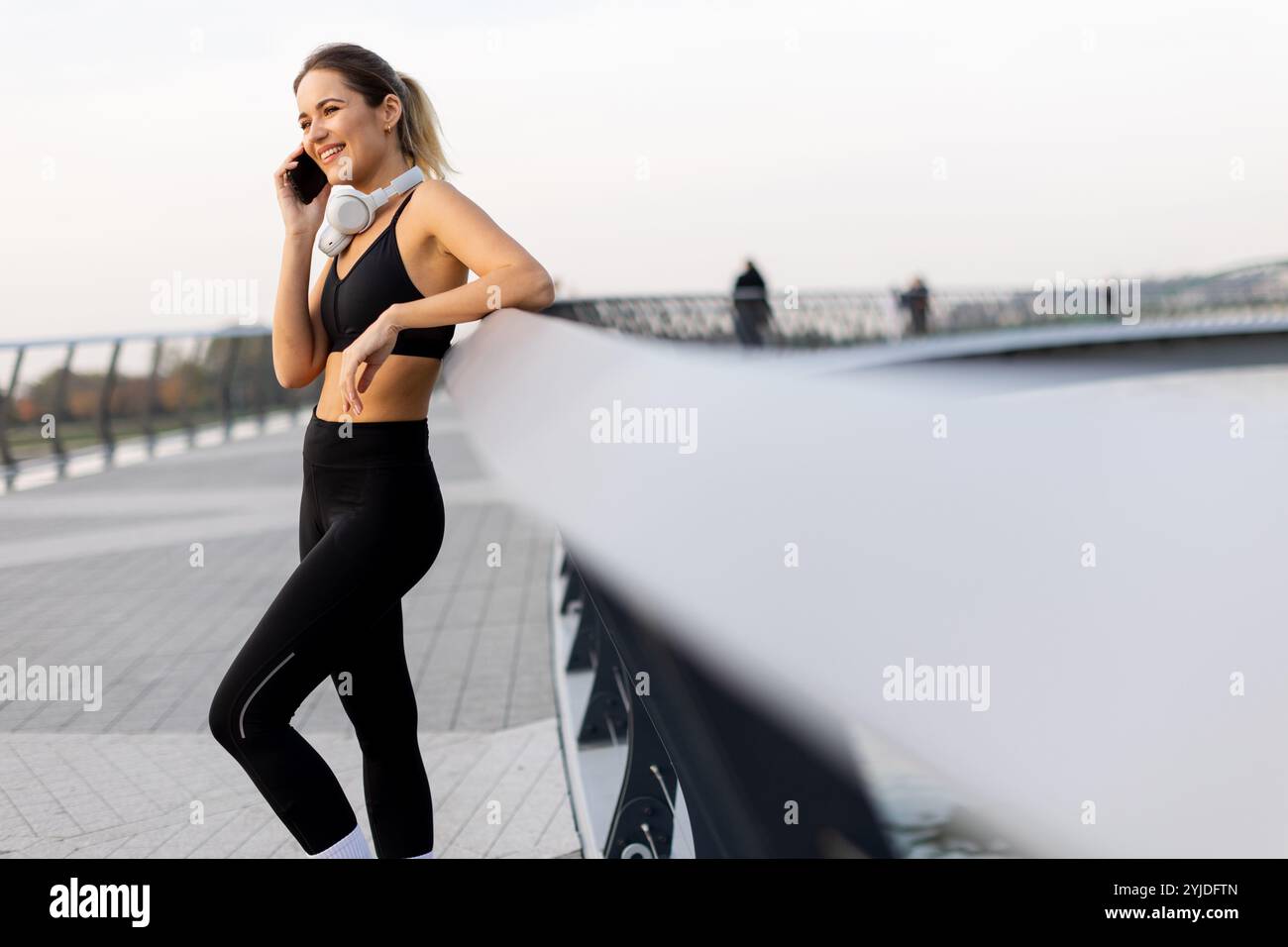 Eine Joggerin hält an ihrem Telefon am Fluss an, trägt Trainingsausrüstung und Kopfhörer und genießt die warme Sonne und die ruhige Atmosphäre Stockfoto