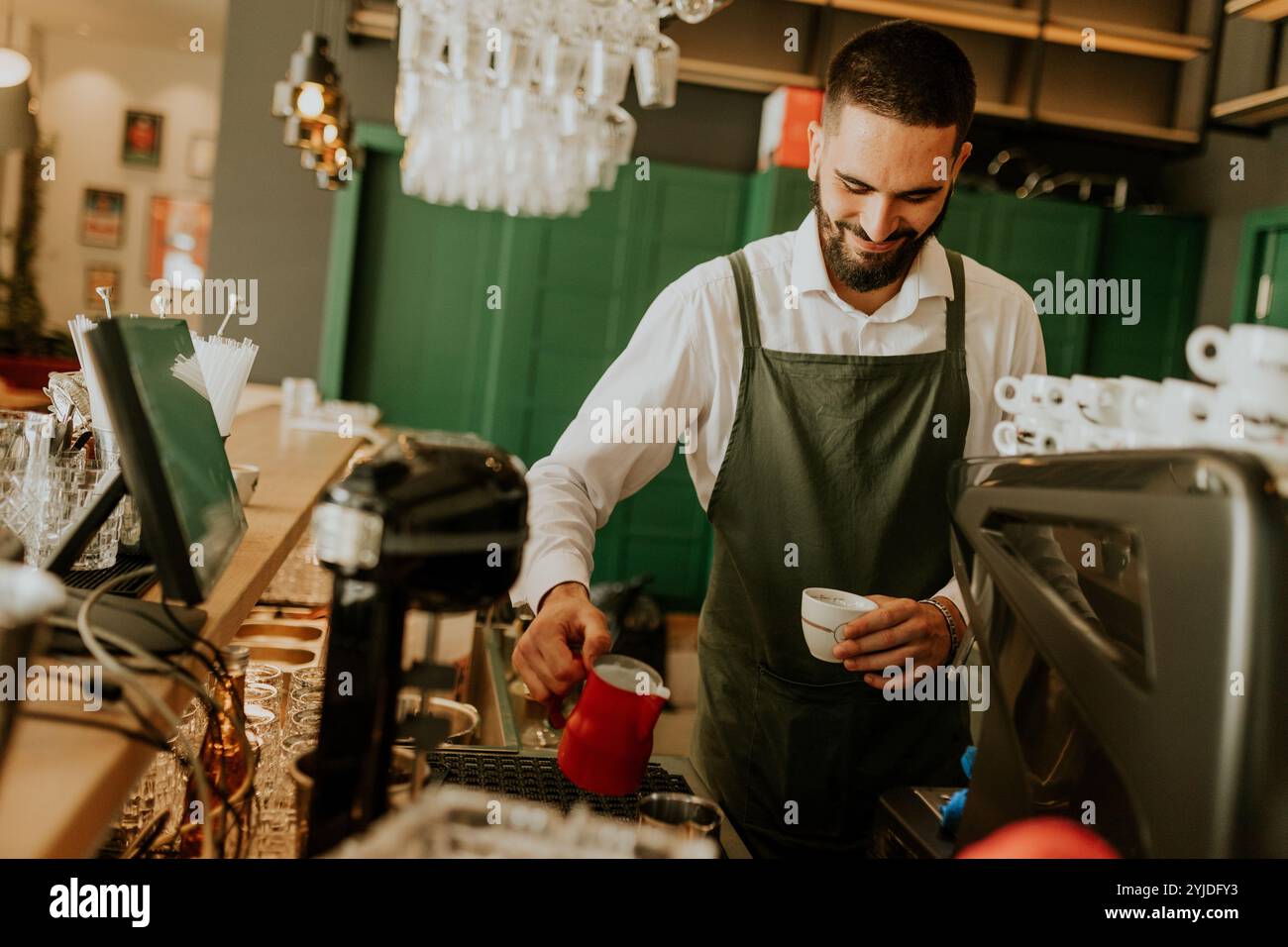 In einem warmen und einladenden Café gießt ein engagierter Barista fachmännisch Milch in eine Kaffeetasse und sorgt so für ein angenehmes Erlebnis für Gäste. Stockfoto
