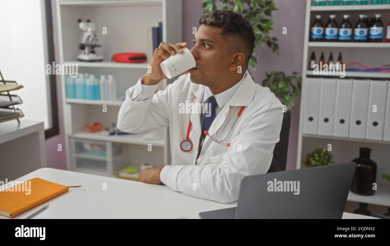 Junger hispanischer Mann trinkt Kaffee am Klinikarbeitsplatz mit weißem Mantel und Stethoskop Stockfoto