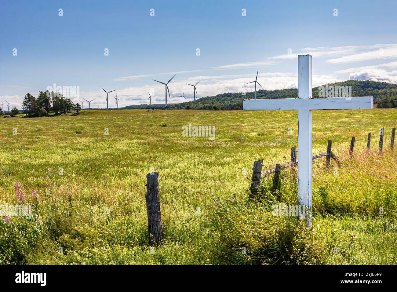 Straßenkreuz in einem Feld mit Gras und Wildblumen, vor Windturbinen. Religiöse Symbole mischen sich mit Technologie. Stockfoto