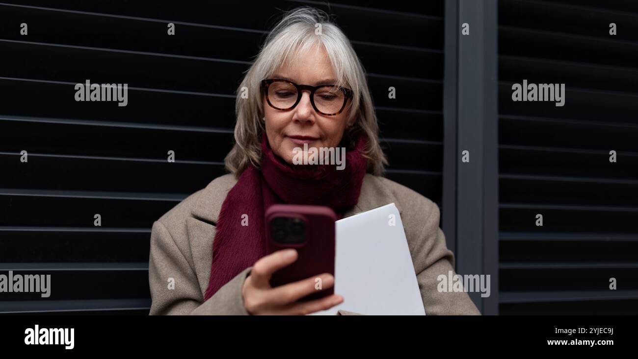 Ältere Geschäftsfrau steht in Winterkleidung vor ihrem Büro, während sie ihr Smartphone während eines geschäftigen Arbeitstages überprüft Stockfoto