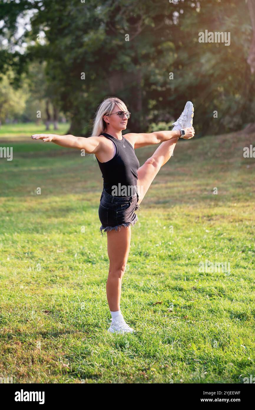 Sportliche Frau, die Utthita Hasta Padangusthasana Nummer eins macht, im Park auf einem Bein mit dem linken Bein oben in lässiger Kleidung, durchgehend VI Stockfoto
