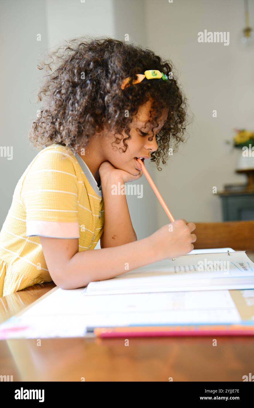 Lächelndes Kind in gelbem Kleid, das mit Bleistift am Schreibtisch studiert. Stockfoto