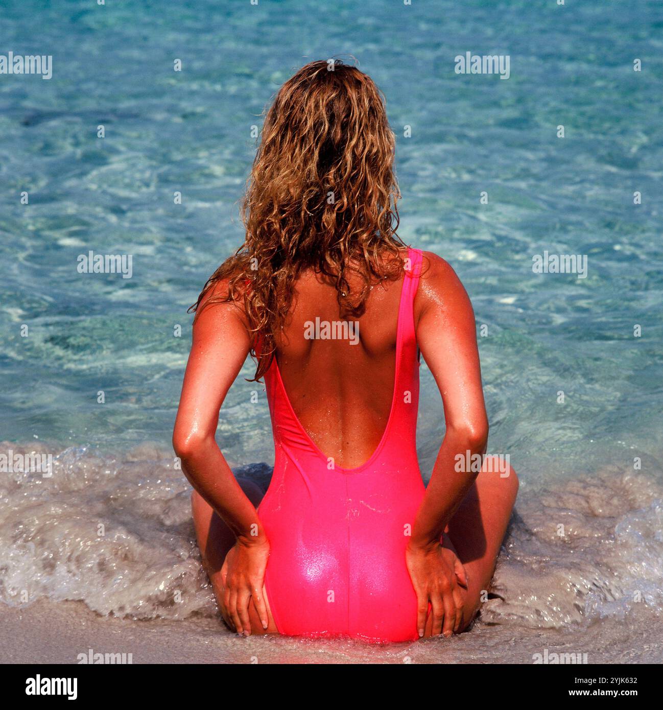 Mittelmeer. Rückansicht einer jungen Frau in rosa Badeanzug, die am Wasserrand sitzt. Stockfoto