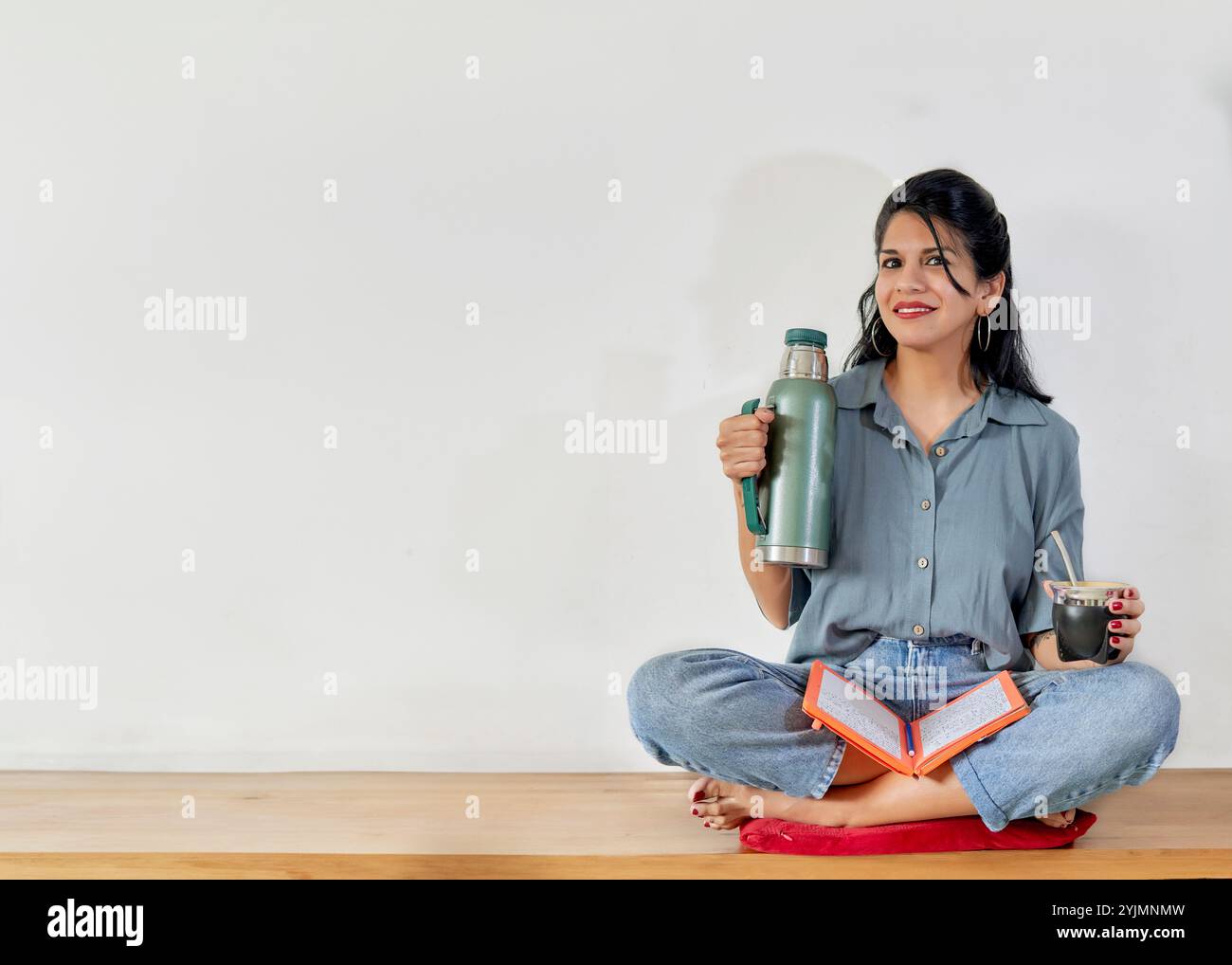 latina sitzt im Kreuz auf einer Holzbank und hält einen Kumpel und eine Thermoskanne, die lächelnd in die Kamera blickt, mit einer Agenda, die ihre Ziele vorsieht. Stockfoto