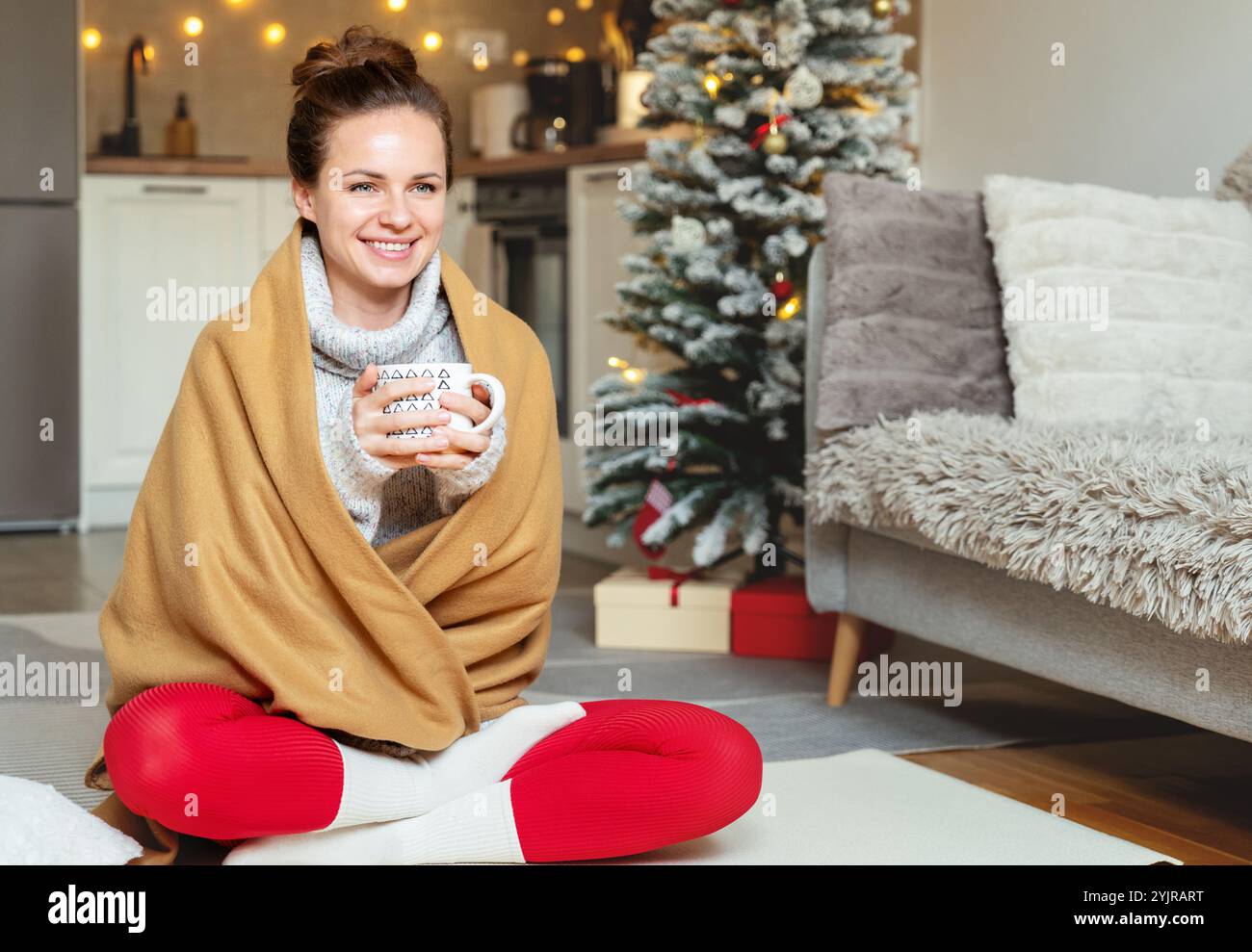 Kaukasische Frau, bedeckt mit einer Fleecedecke, sitzt zu Hause auf dem Boden und trinkt während Weihnachten ein heißes Getränk aus einer Tasse Stockfoto