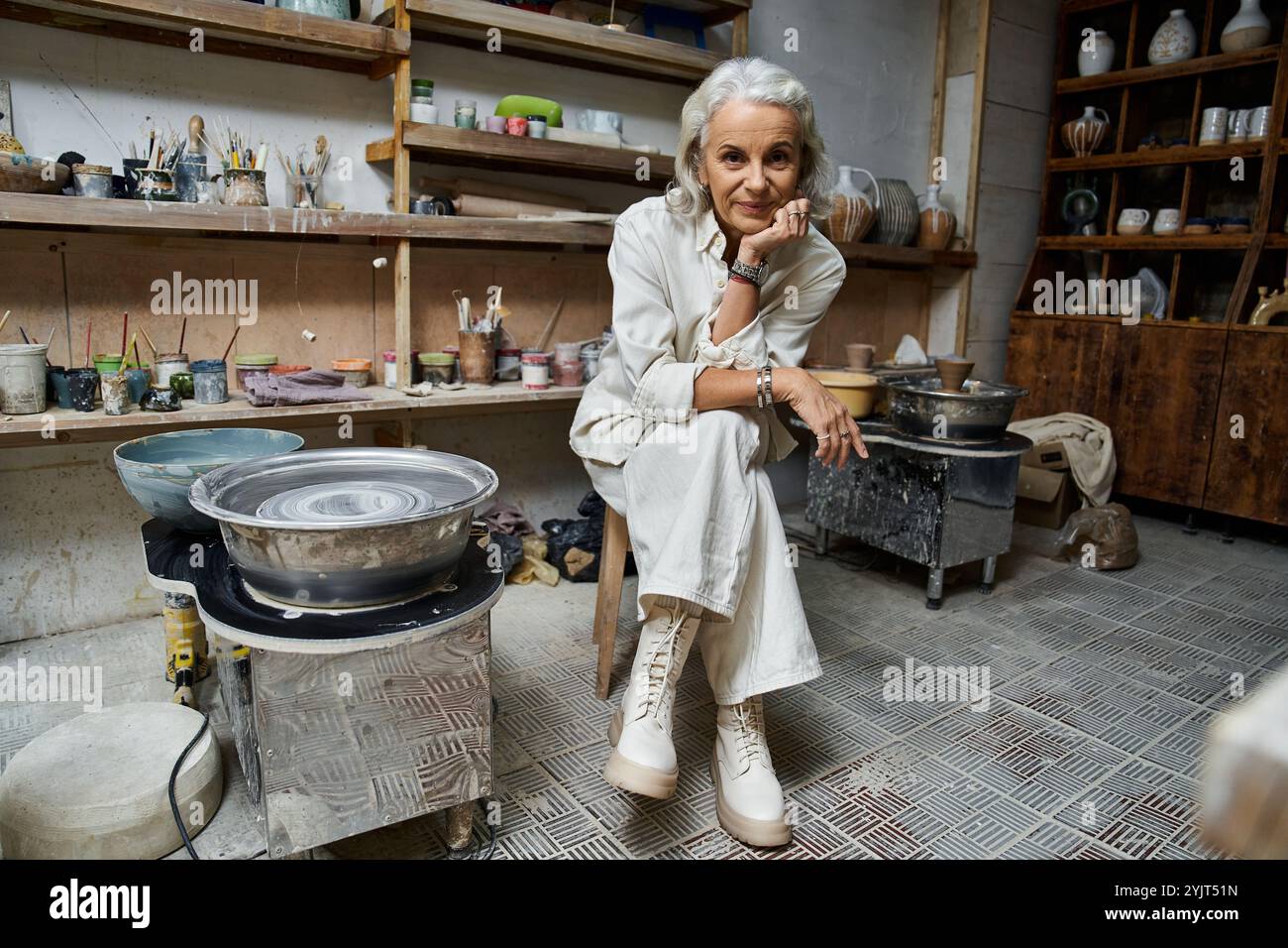 Eine erfahrene Handwerkerin arbeitet in Töpfertechniken, während sie von ihrem kreativen Arbeitsbereich umgeben ist. Stockfoto