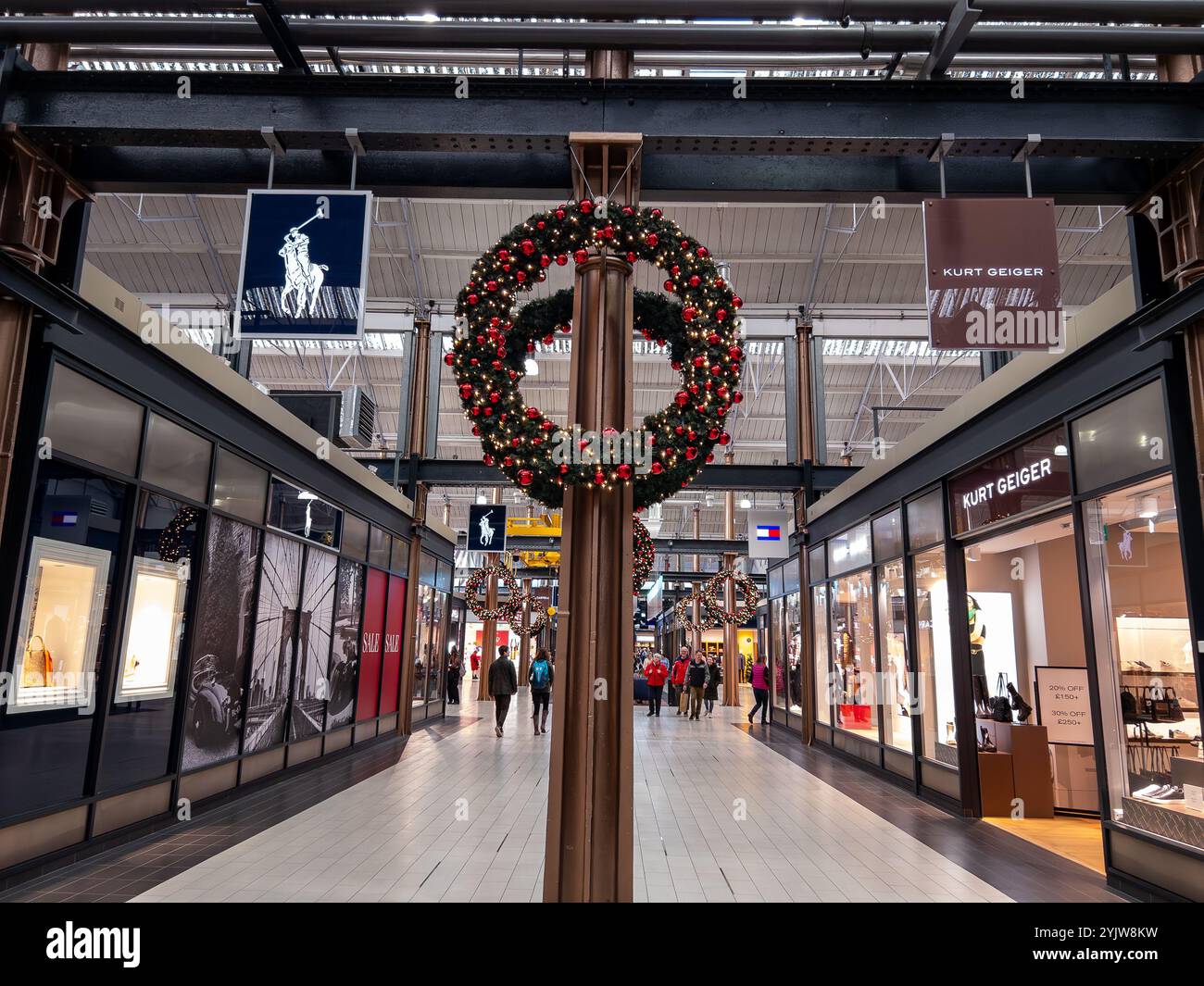 Der festliche Weihnachtskranz schmückt den Gang eines Einkaufszentrums, während die Leute während der Weihnachtszeit an Ladengeschäften im Swindon Outlet Center 2024 vorbeischlendern - Smartphone-aufgenommenes Stockfoto