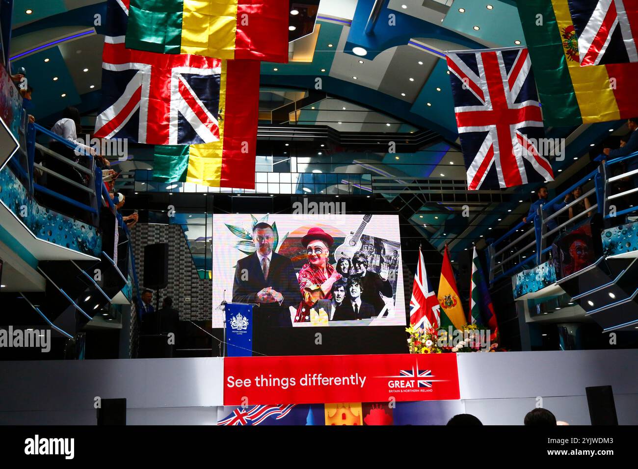 El Alto, BOLIVIEN; 15. November 2024: Ein Bild des britischen Botschafters Richard Porter, eines traditionellen bolivianischen Diablada-Tänzers, und der Beatles, die auf einer Großleinwand, umgeben von Union Jacks und bolivianischen Flaggen, zu Beginn einer Veranstaltung der britischen Botschaft in Bolivien anlässlich des Geburtstages von König Karl III. (Der gestern war) gezeigt wurden. Die Veranstaltung sollte auch die kulturellen Beziehungen und den Handel zwischen Bolivien und Großbritannien fördern. Stockfoto