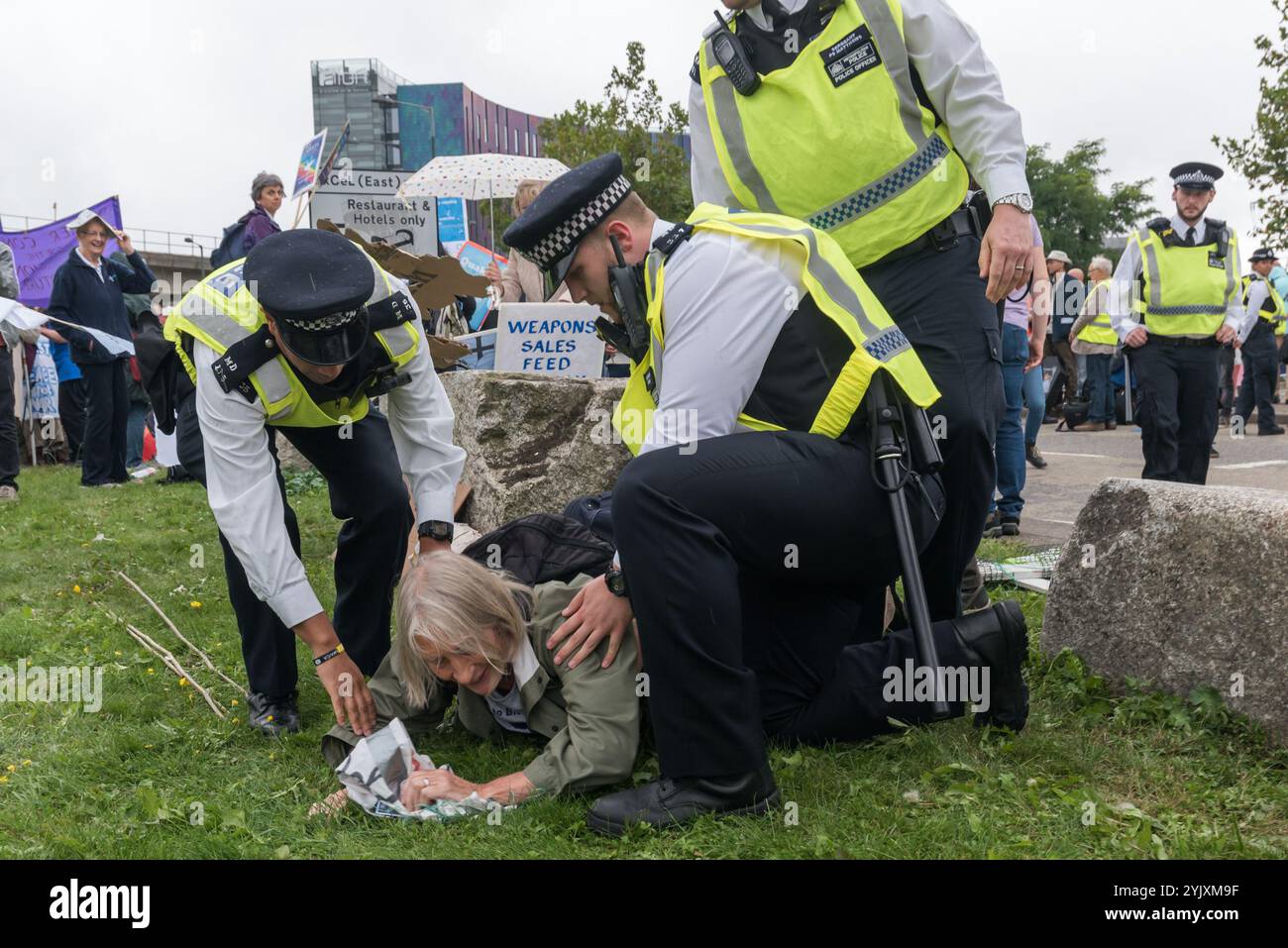 London, Großbritannien. 5. September 2017. Polizei vorsichtig ein Quäker demonstrant sie weg von der Straße auf das Gras am zweiten Tag der Proteste gegen die größten Waffen der Welt in den Londoner Docklands statt durchgeführt haben. Die "Kein Vertrauen in Krieg" Tag war eine Reihe von Veranstaltungen, die von verschiedenen Glauben Gruppen organisiert. Bevor ich ankam hatte es einen Lock-in auf der Zufahrtsstraße stoppen Lieferungen auf der Messe über das Tor zu setzen. Dies wurde gefolgt von einem Quaker Meeting an der Seite der Straße, bei dem eine Reihe von Personen stand oder lag die Straße zu blockieren und mehrere, die sich weigerten, sich zu bewegen, wurden festgenommen. Stockfoto