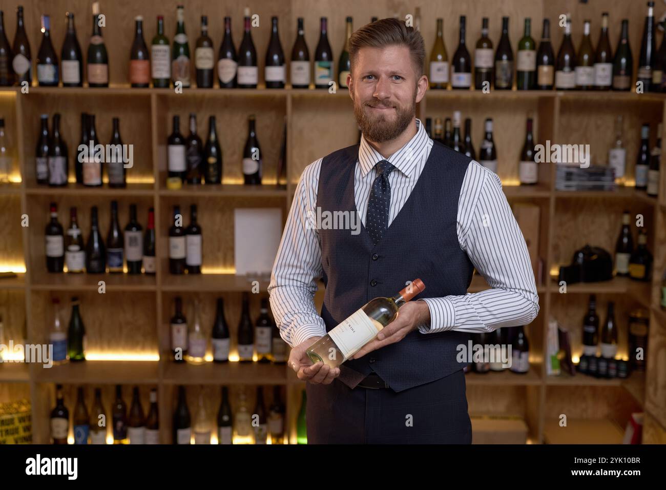 Porträt eines lächelnden professionellen Sommeliers, der eine teure Flasche Wein hält, während er im Weinkeller mit Holzregalen für Flaschen posiert Stockfoto