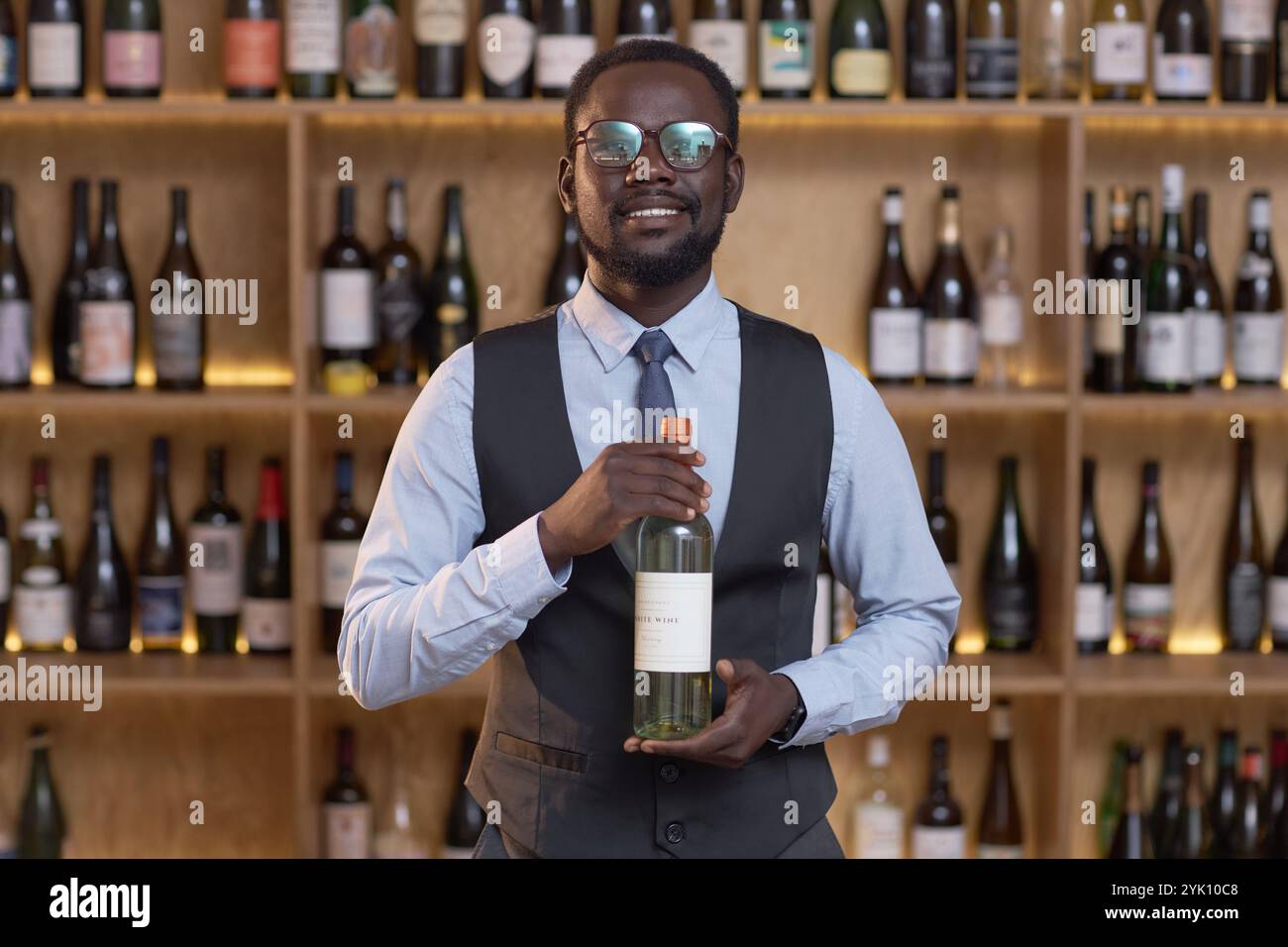 Porträt eines afroamerikanischen Sommeliers, der weit lächelt, während er eine Flasche Weißwein stolz im Weinkeller hält, Holzhütten mit einer riesigen Anzahl von Weinflaschen im Hintergrund Stockfoto