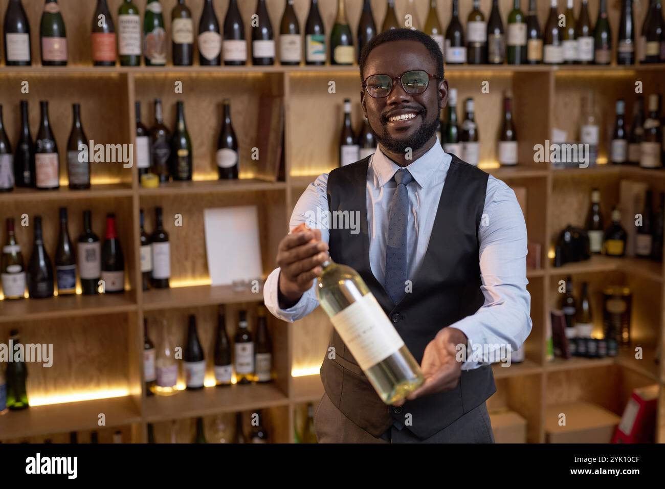 Lächelnder afroamerikanischer Sommelier in eleganter Kleidung, der in die Kamera blickt, während er exklusiven Wein vorführt Stockfoto