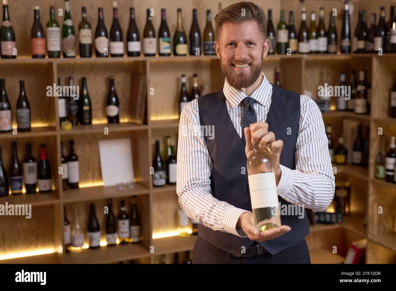 Porträt eines strahlenden kaukasischen Sommeliers mit einer Weinflasche, die im Weinkeller zum Foto posiert Stockfoto