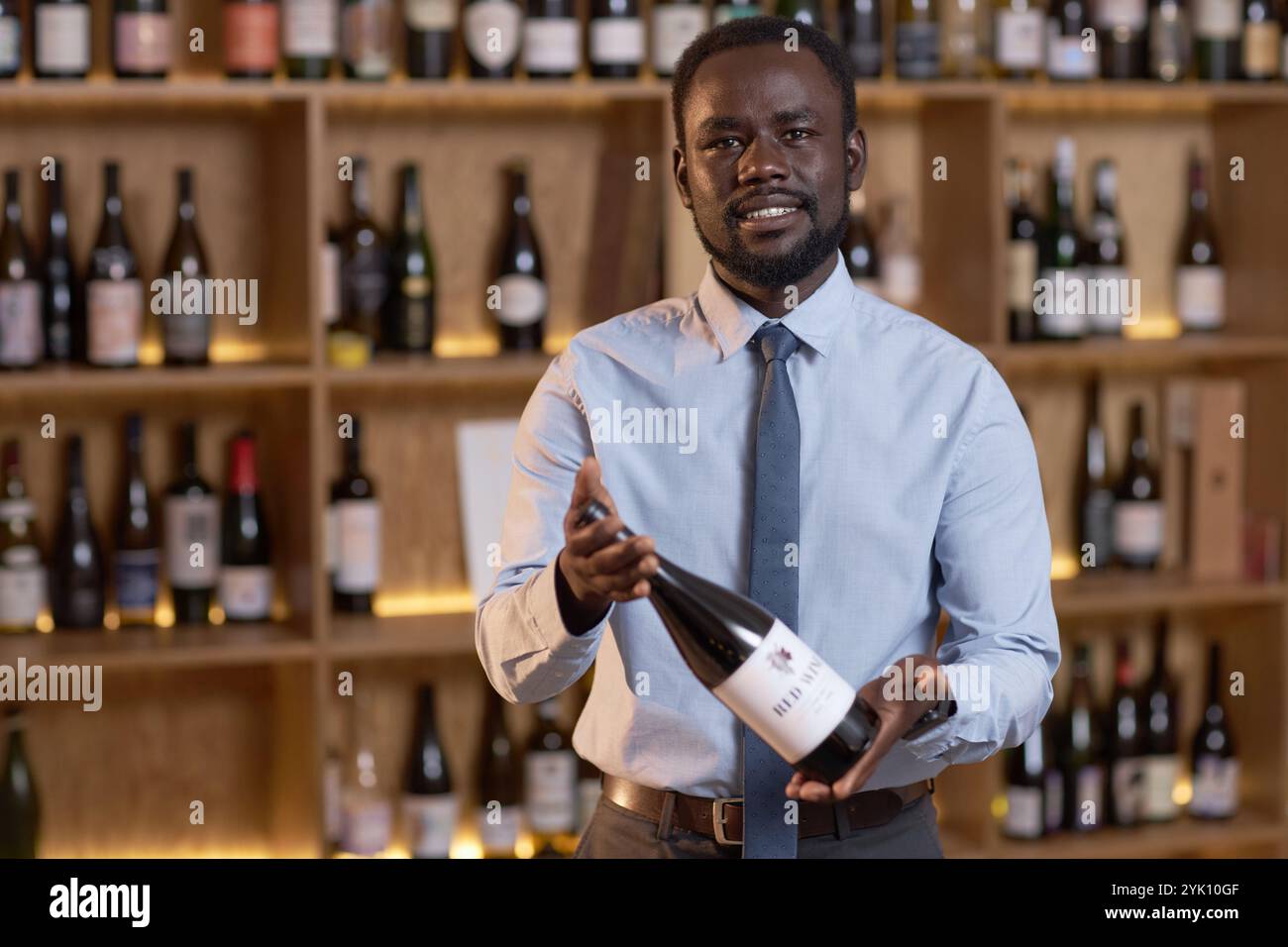 Mittelgroßes Nahporträt eines afroamerikanischen Sommeliers, der eine Rotweinflasche hält, während er für ein Foto im Weinkeller voller Wein posiert Stockfoto