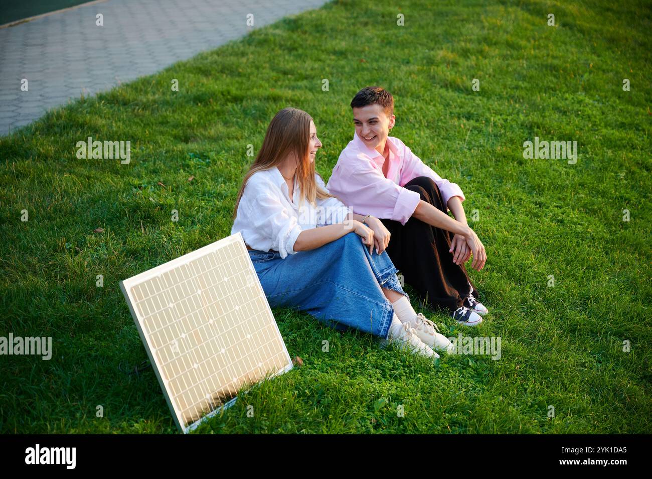 Junge Frauen, die Solarpanel zum Laden verwenden, sitzen auf grasbewachsenem Rasen, lächeln und reden, mit Solarpanel in der Nähe. Mädchen, die locker gekleidet sind, genießen sonnige, entspannte Momente im Freien. Stockfoto