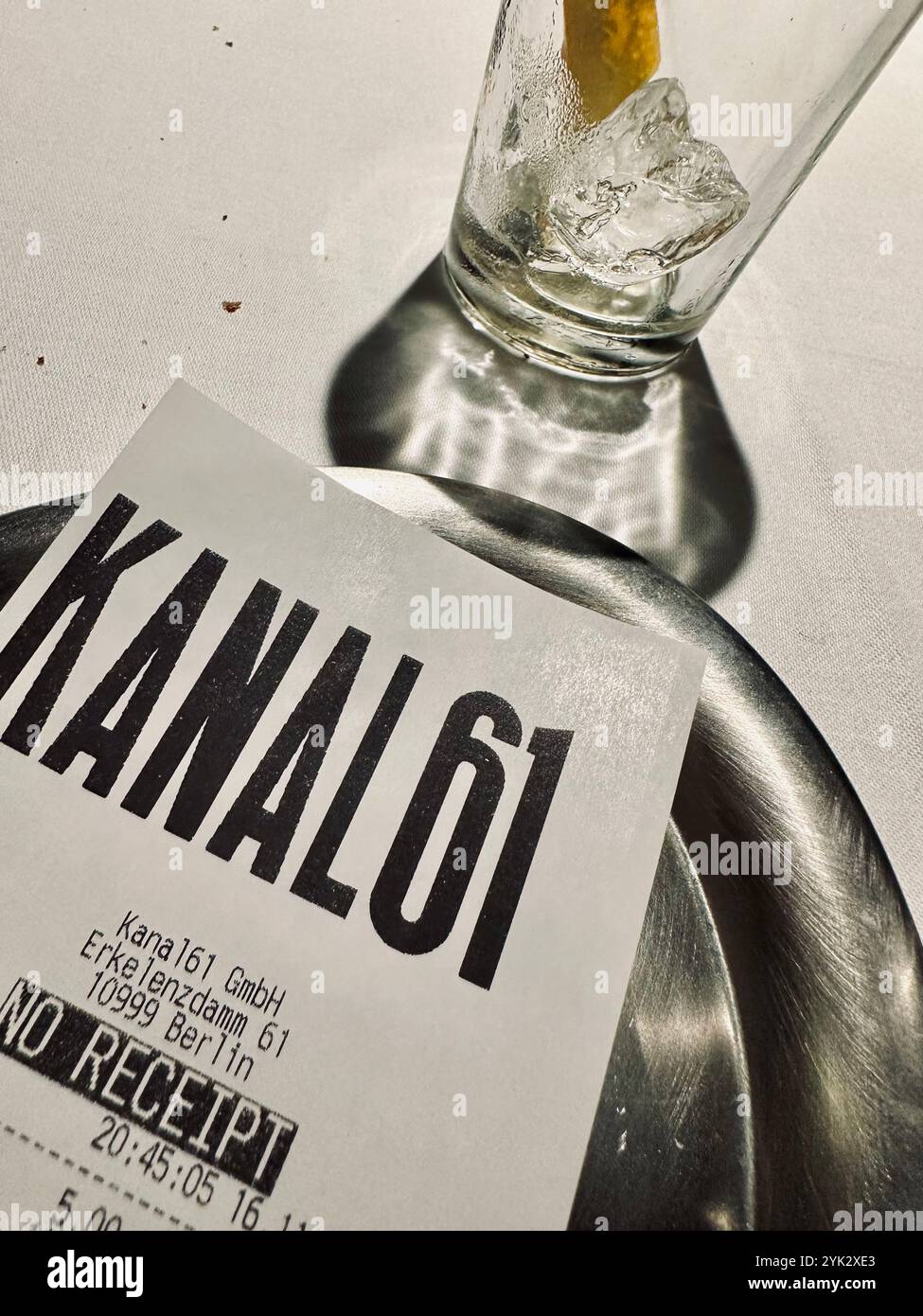 Eine Rechnung des beliebten Berliner Restaurants Kanal61 auf einem kleinen silbernen Tablett auf weißer Tischdecke mit einem leeren Glas mit Eiswürfeln im Hintergrund - Smartphone-aufgenommenes Stockfoto