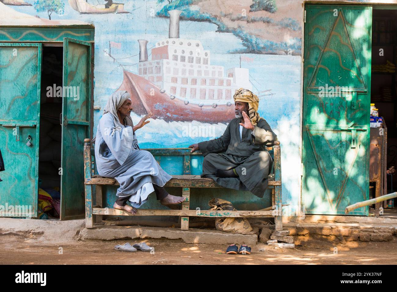 Ältere Männer sitzen auf einer Bank unter einem Wandgemälde in einer Straße des Dorfes Ramadi, Westufer des Nils südlich von Edfu, Ägypten, Nordostafrika Stockfoto