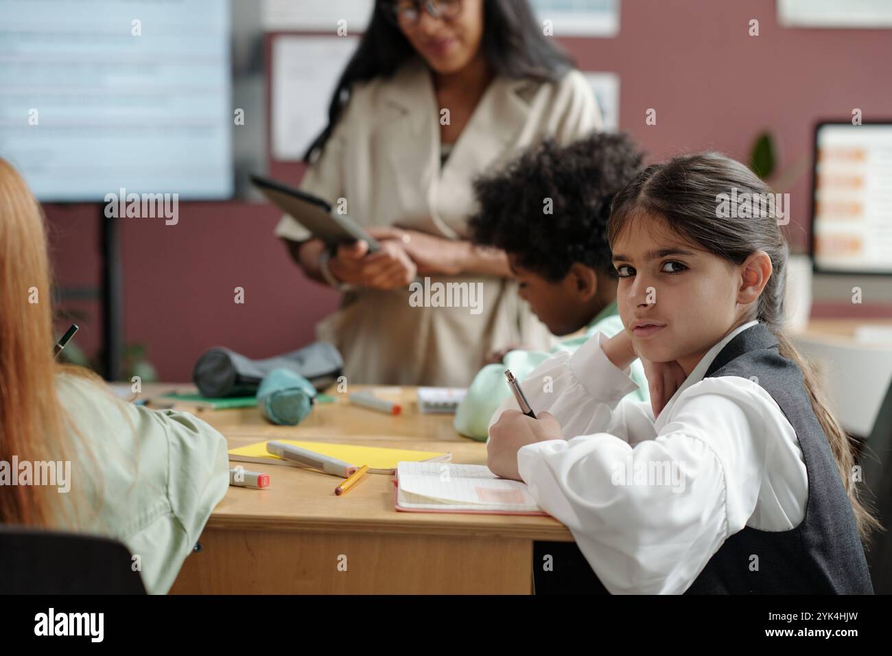 Ernsthaftes Grundschulmädchen, das in die Kamera schaut und Notizen im Copybook macht, während es am Schreibtisch beim Englischunterricht sitzt Stockfoto