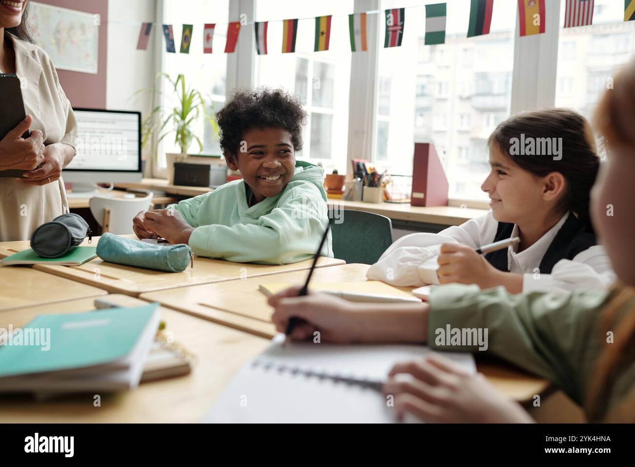 Glücklicher, süßer Schuljunge im Hoodie, der beim Unterricht am Schreibtisch sitzt und einen der Klassenkameraden während der Diskussion der Grammatikregeln ansieht Stockfoto