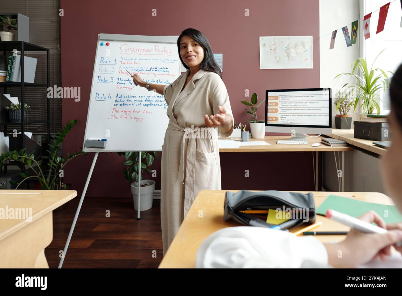 Glückliche junge Lehrerin in lässigem Kleid, die auf das Whiteboard zeigt, mit Grammatikregeln und auf das Schulmädchen im Unterricht schaut Stockfoto