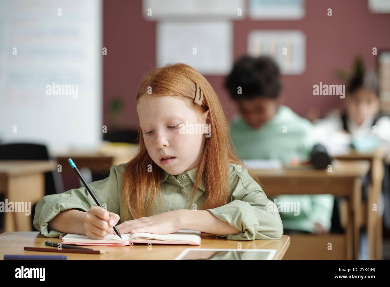Kluges und fleißiges Schulmädchen, das Notizen auf der Seite des Copybook macht, während es am Schreibtisch im Klassenzimmer sitzt und Englischgrammatikunterricht erhält Stockfoto