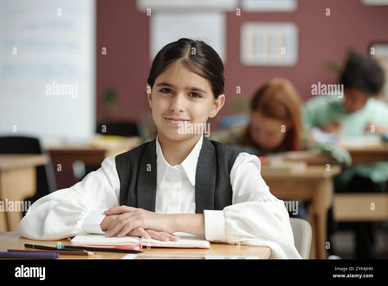 Fleißige brünette Schulmädchen sitzt am Schreibtisch, während sie Englisch-Grammatik lernt und ihre Klassenkameraden in die Kamera sieht Stockfoto