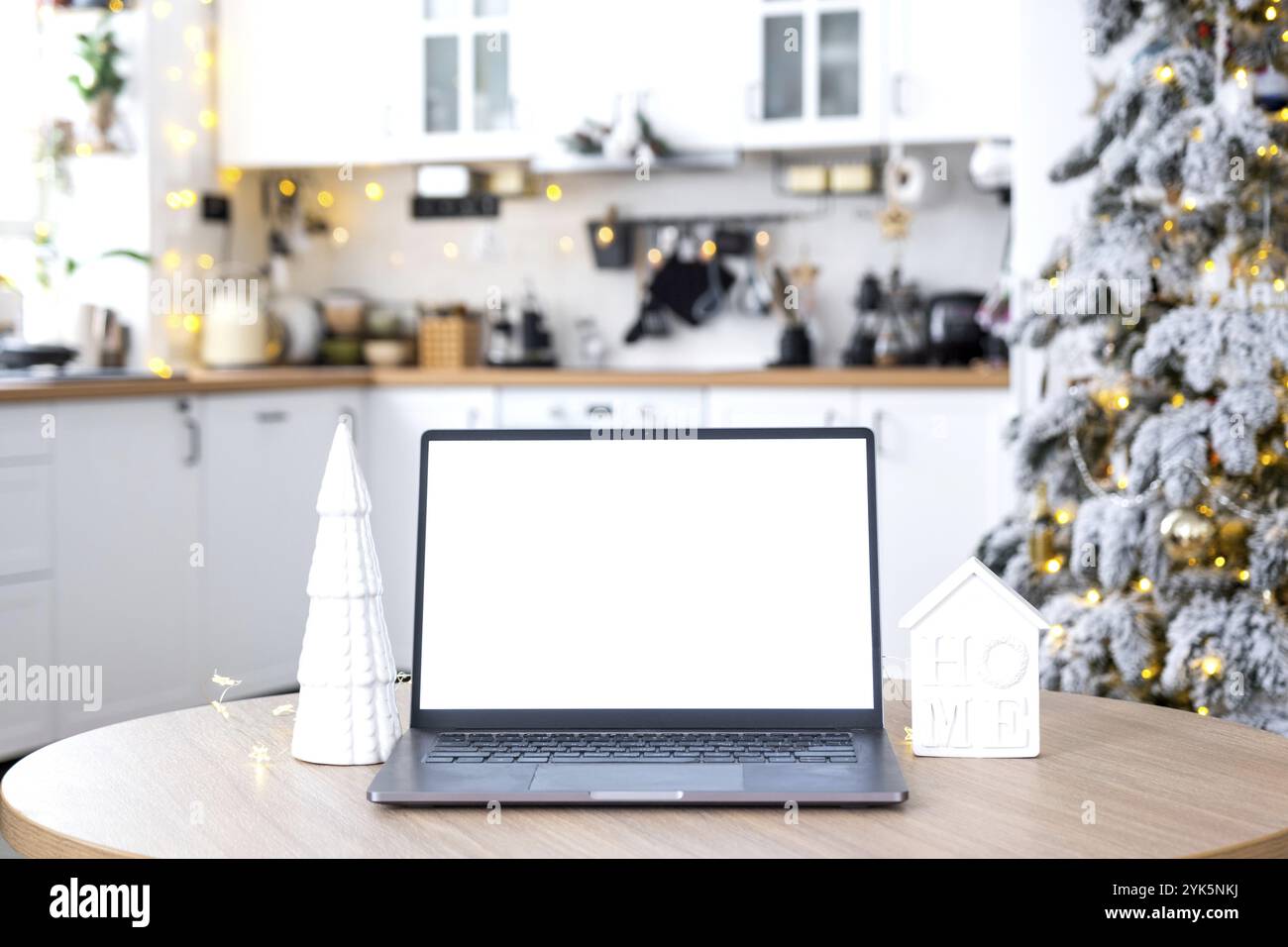 Laptop mit einem weißen Bildschirm in der gemütlichen, weiß dekorierten Weihnachtsküche mit Feenlicht und einem Weihnachtsbaum. Saisonale Fernarbeit, intern Stockfoto