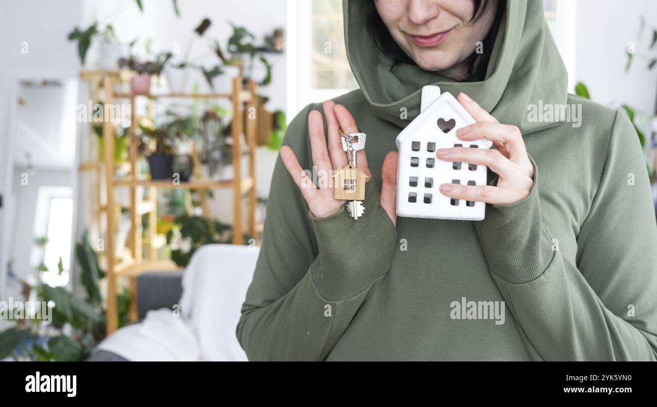 Eine glückliche Frau in ihrem Haus hält in ihren Händen eine Miniaturfigur eines Hauses und einen Schlüssel im Inneren. Traumhausprojekt, Immobilienkauf, Versicherung Stockfoto