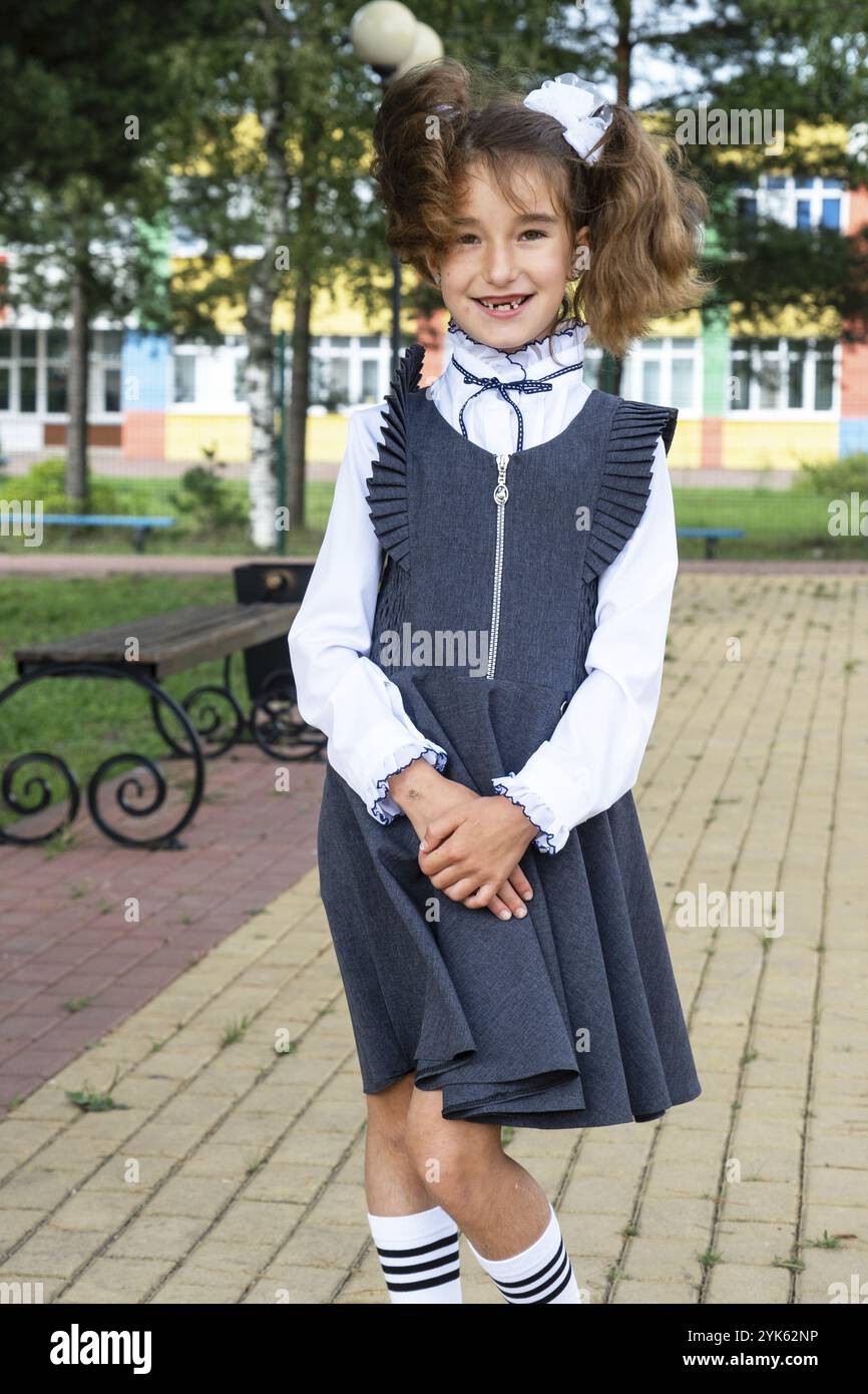 Fröhliches, lustiges Mädchen mit einem zahnlosen Lächeln in einer Schuluniform mit weißen Bögen im Schulhof. Zurück zur Schule, 1. September. Ein glücklicher Schüler. Primäres edu Stockfoto