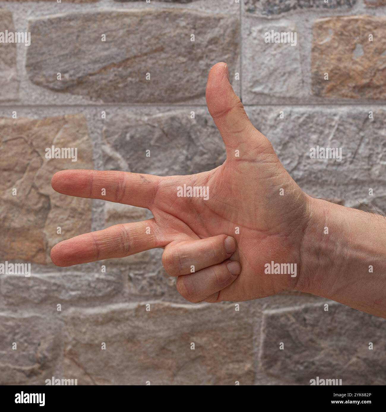 Hand, die eine Geste mit drei Fingern vor einer strukturierten Steinmauer macht Stockfoto