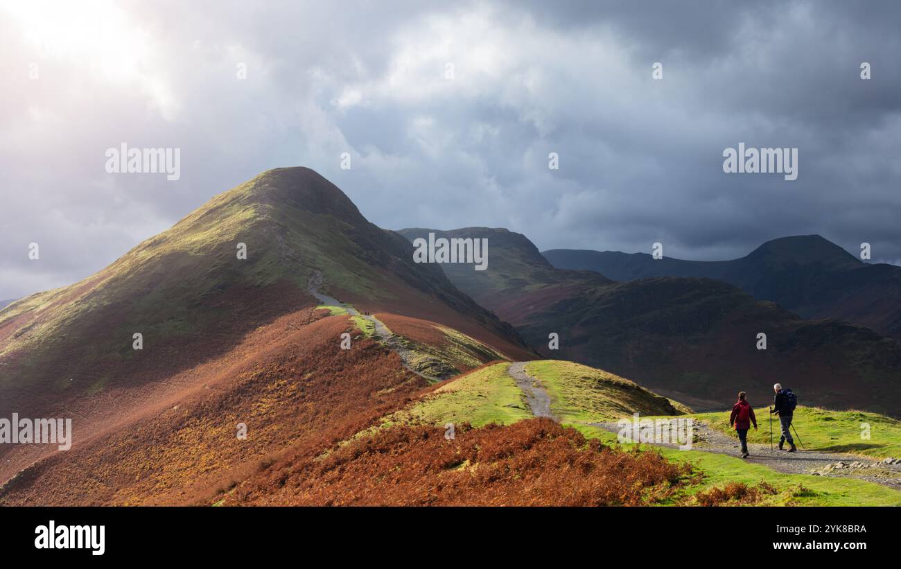 Wanderer, die den Pfad hinauf auf Catbells im Lake District, Cumbria, England, mit umliegenden Fjells unter dramatischen Wolken. Stockfoto