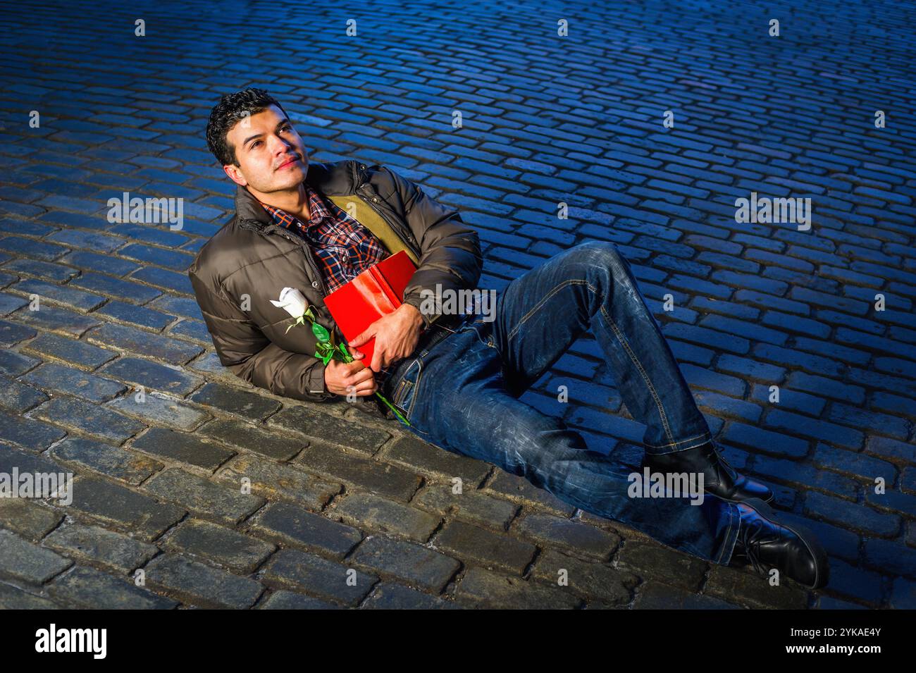 Ein Mann liegt auf einer kopfsteingepflasterten Straße, sieht besinnlich aus, während er ein rotes Buch und eine weiße Rose hält. Er trägt eine lässige Jacke und Jeans Stockfoto