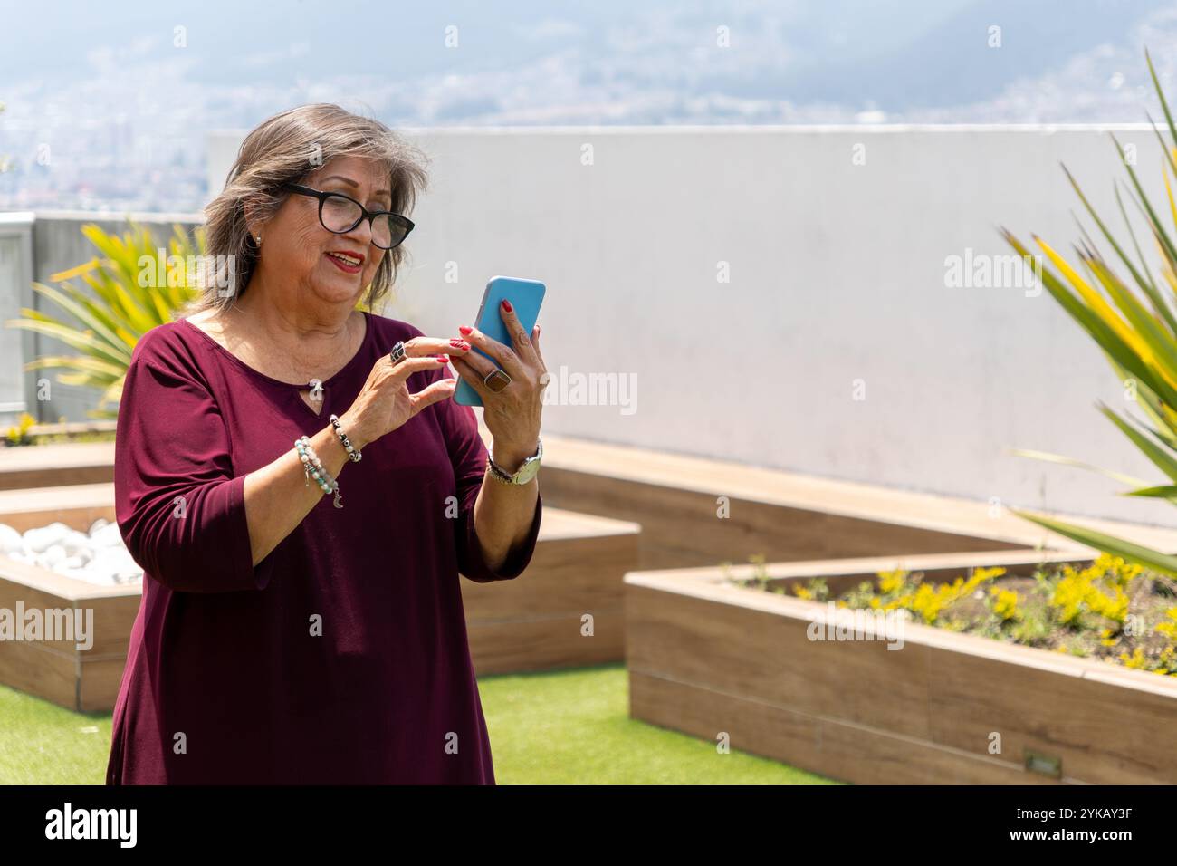 Senior Latina Frau, die morgens im Garten lächelnd auf ihr Handy blickt Stockfoto
