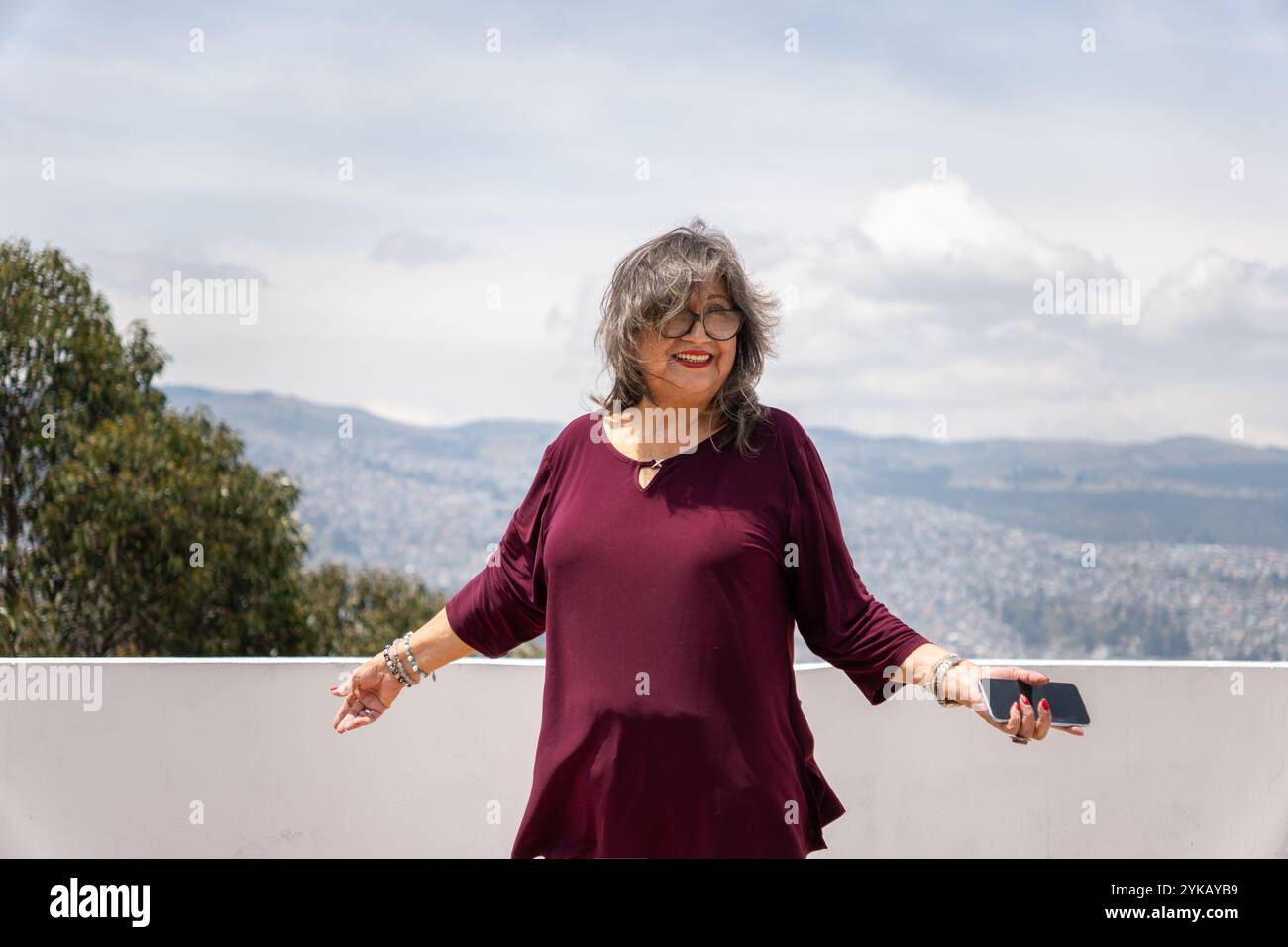 Glückliche Seniorin lächelt mit ihrem Handy in der Hand, während der Wind ihr Haar auf einer Terrasse an einem Sommermorgen bewegt Stockfoto