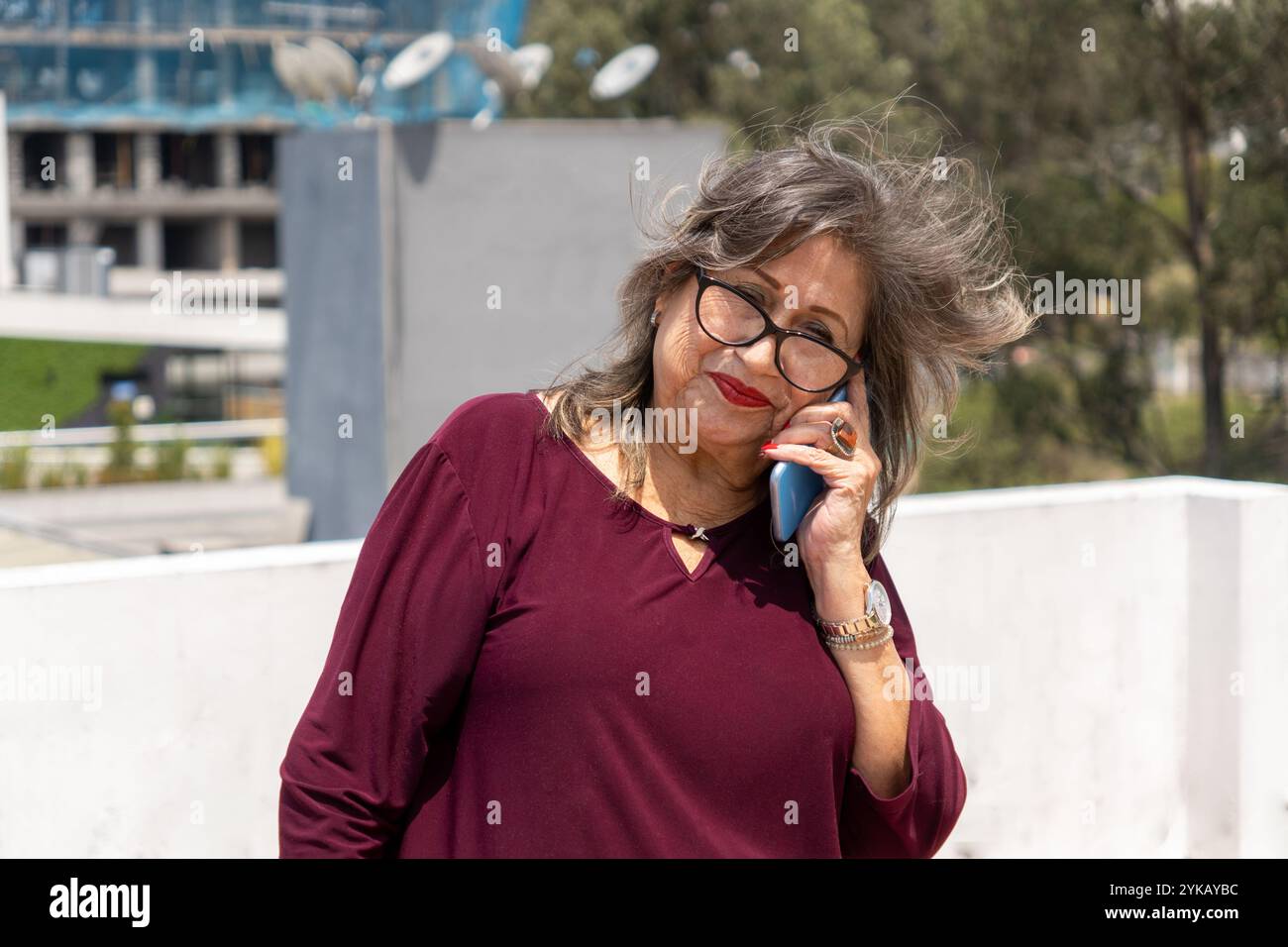 Senior Latina Frau mit Brille, die am windigen Morgen auf einer Terrasse mit Bäumen und Gebäuden spricht Stockfoto